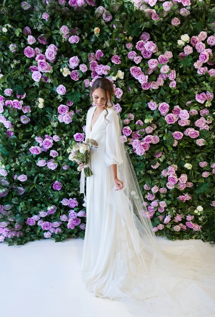 A bride in a white wedding dress holding a bouquet of flowers, standing in front of a lush floral wall of pink and purple roses.