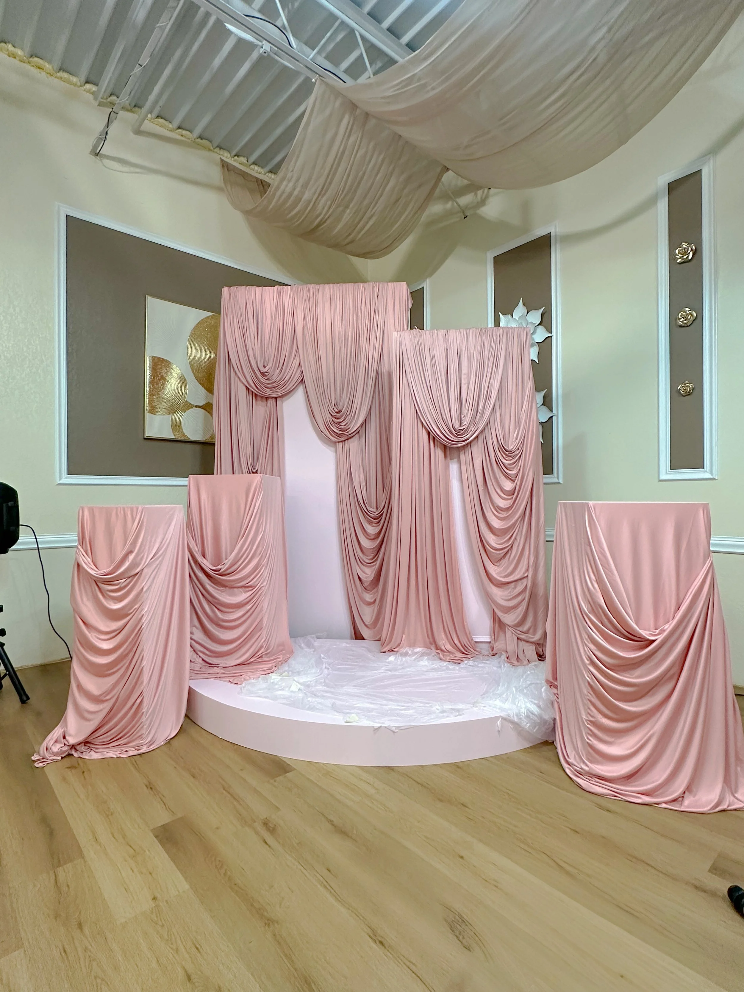 A stage decorated with pink draped fabric, with a small platform and white floor covering, set against a wall with gold and white art and beige panels, in preparation for an event or celebration.