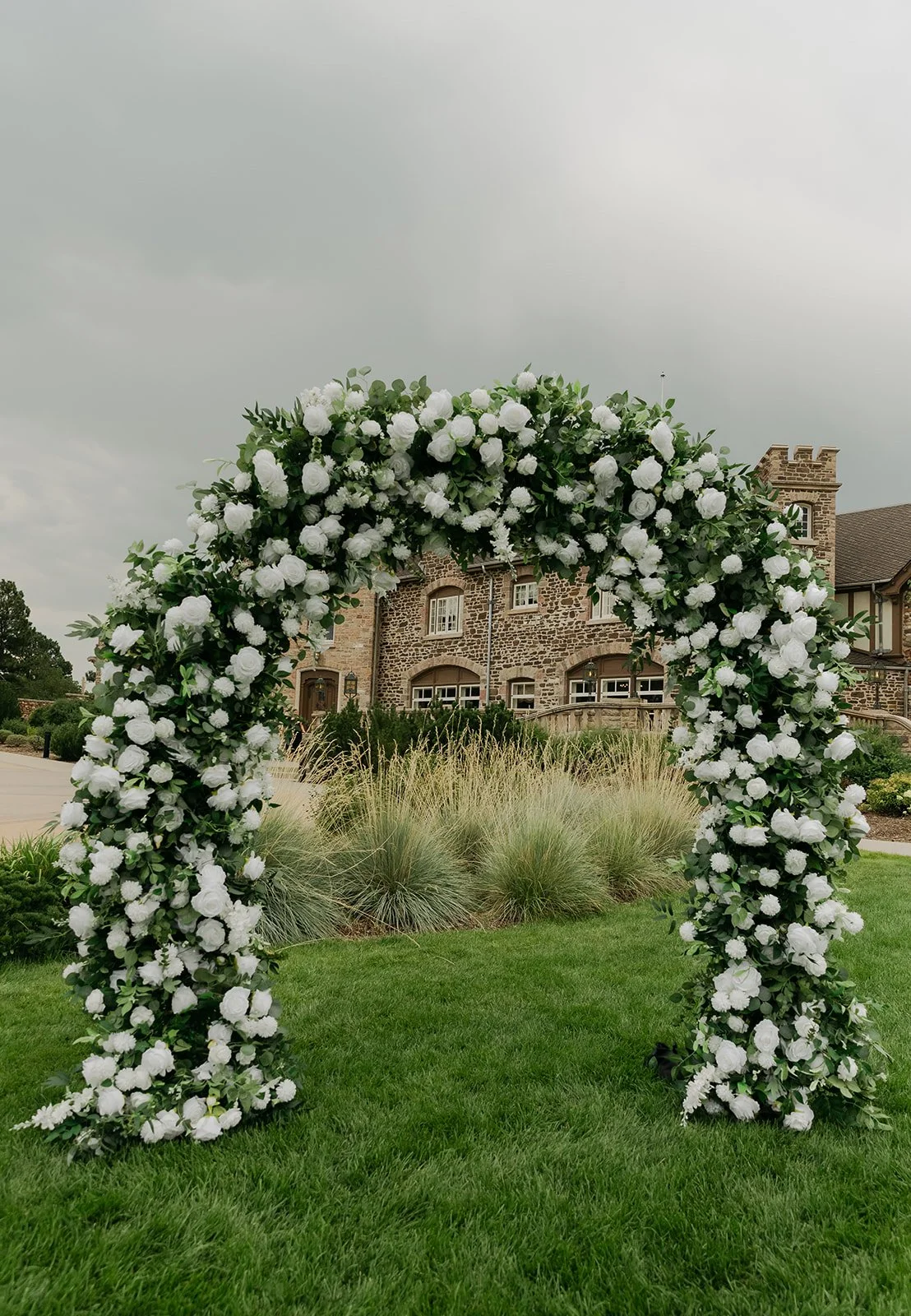 A floral arch with white flowers and green leaves on a lawn, with a brick house in the background under a cloudy sky.