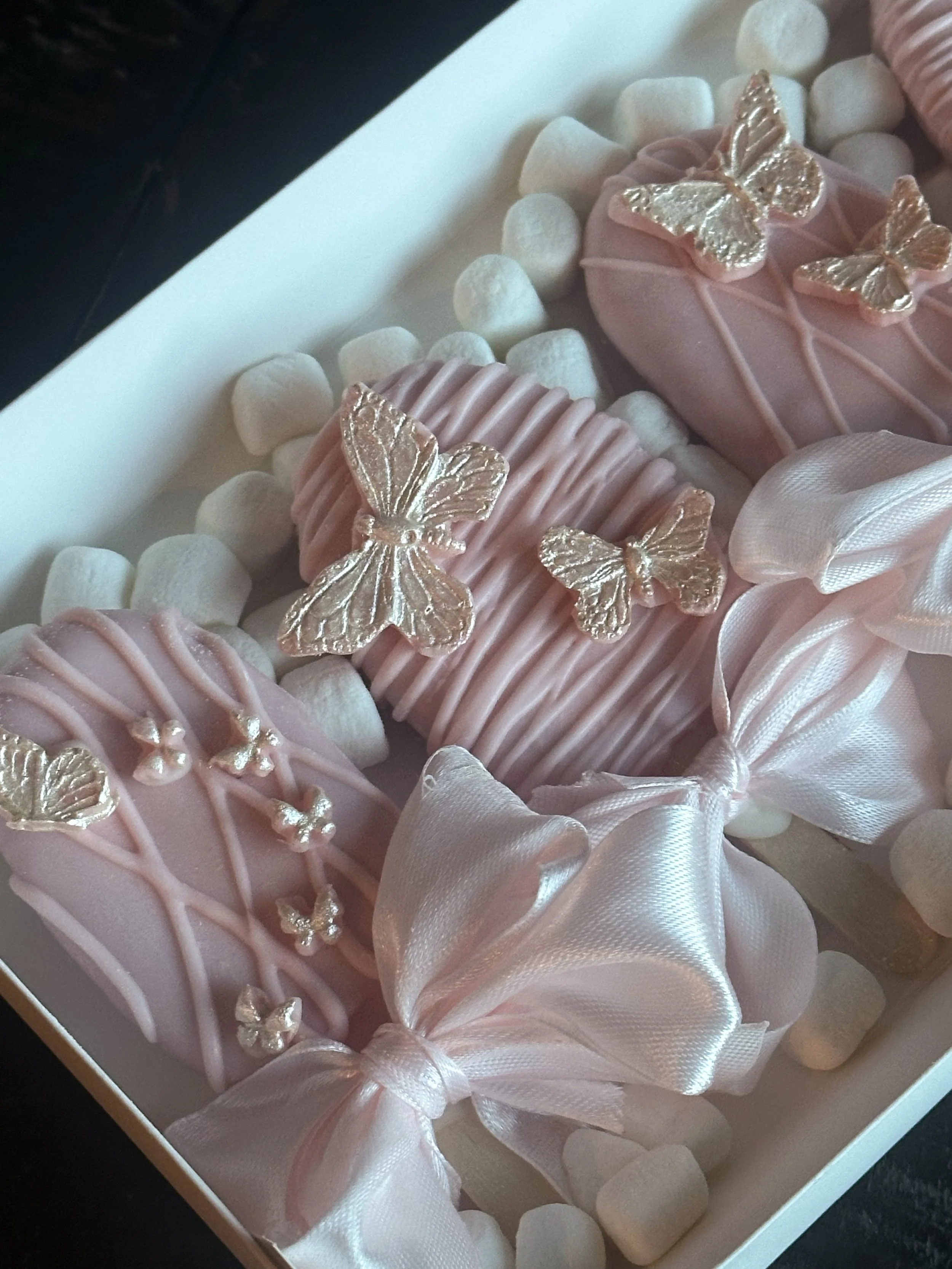 Pink cakepops decorated with silver butterflies and bows, placed in a box with marshmallows.