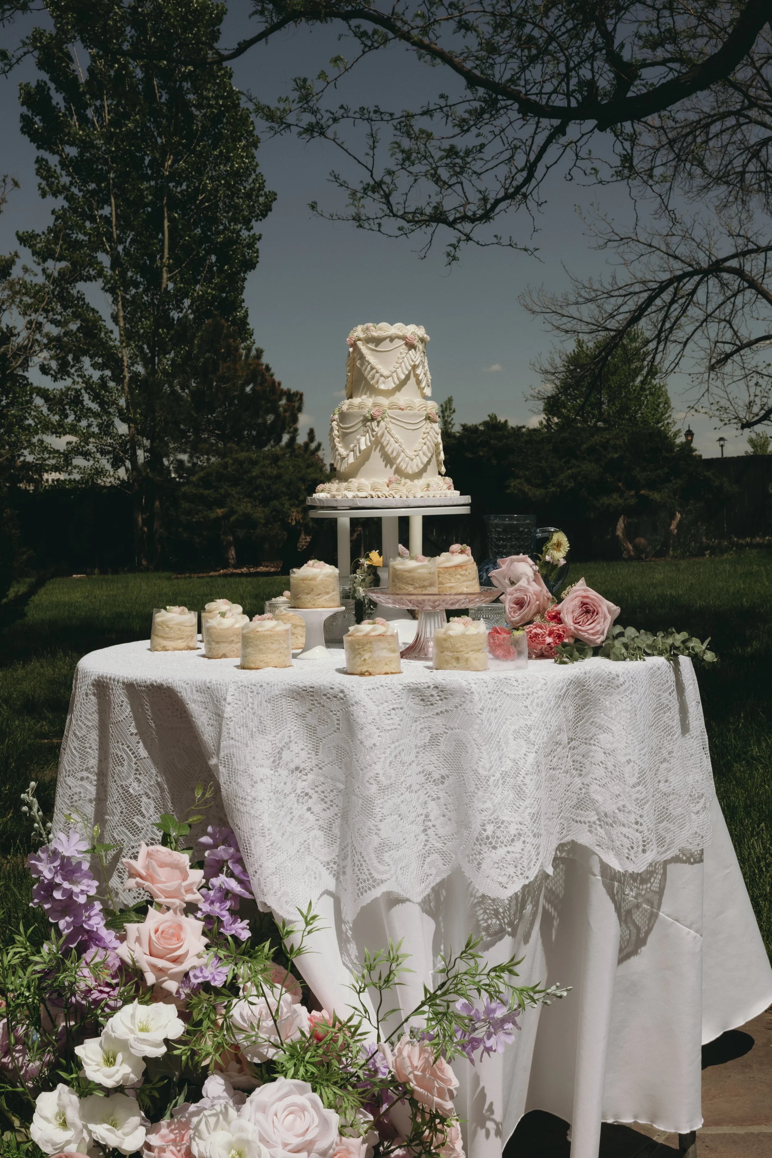 Decorated outdoor table with a large tiered wedding cake, smaller cakes, and floral arrangements against a background of trees and a partly cloudy sky.