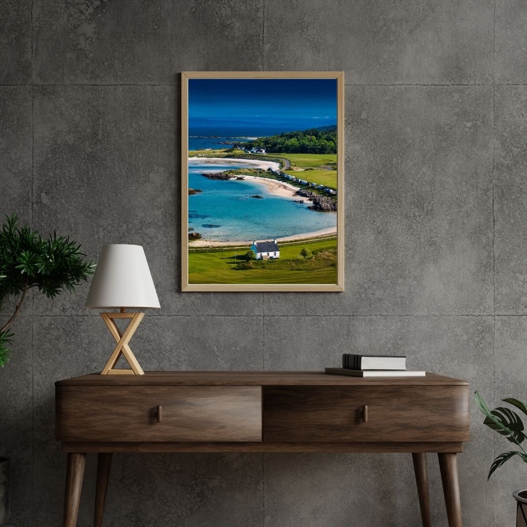 A modern interior scene featuring a framed fine art photograph by James Alroca of Traigh Beach, Scotland. The image captures a coastal scene with a beach, turquoise water, green dunes, and a small white house. The framed print hangs on a concrete wal