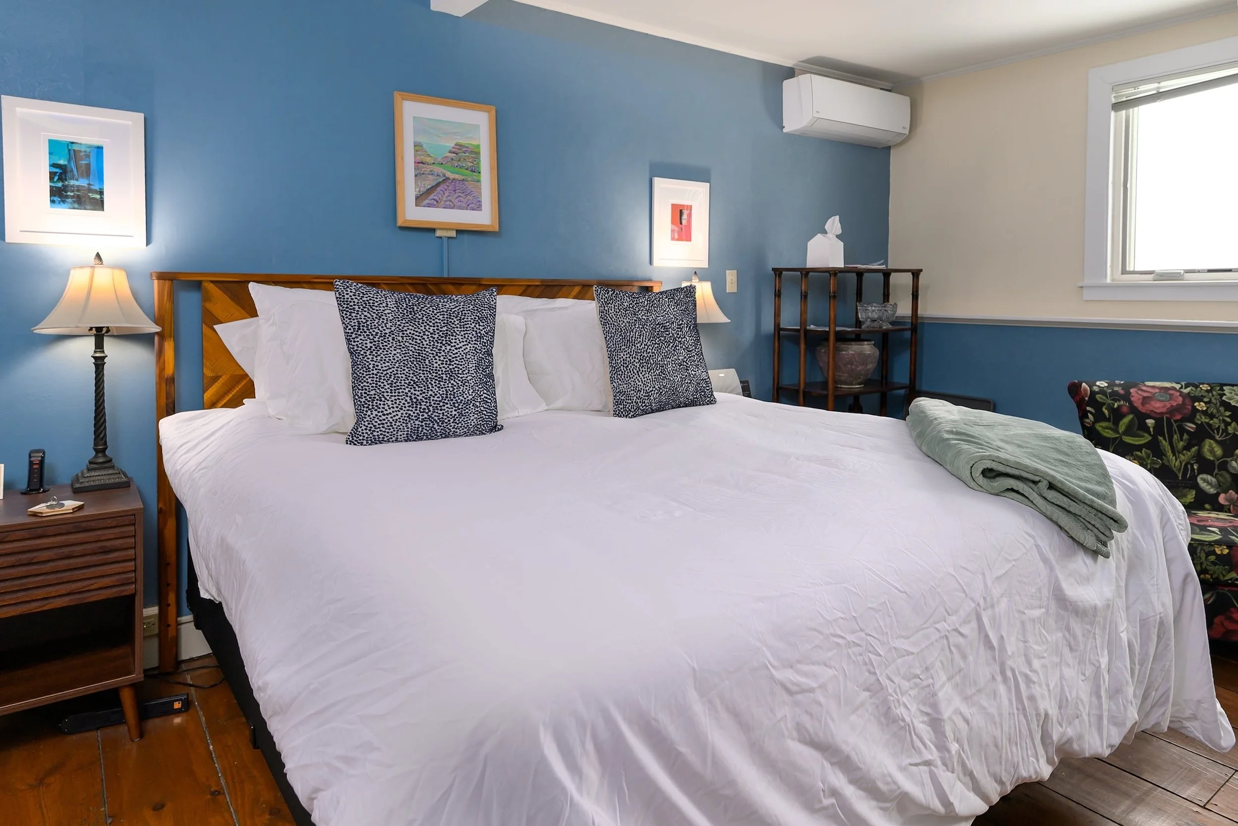 Hotel bedroom with a large bed, nightstands with lamps, a dresser with a TV, and artwork on the walls, featuring hardwood floors and a window with beige curtains.