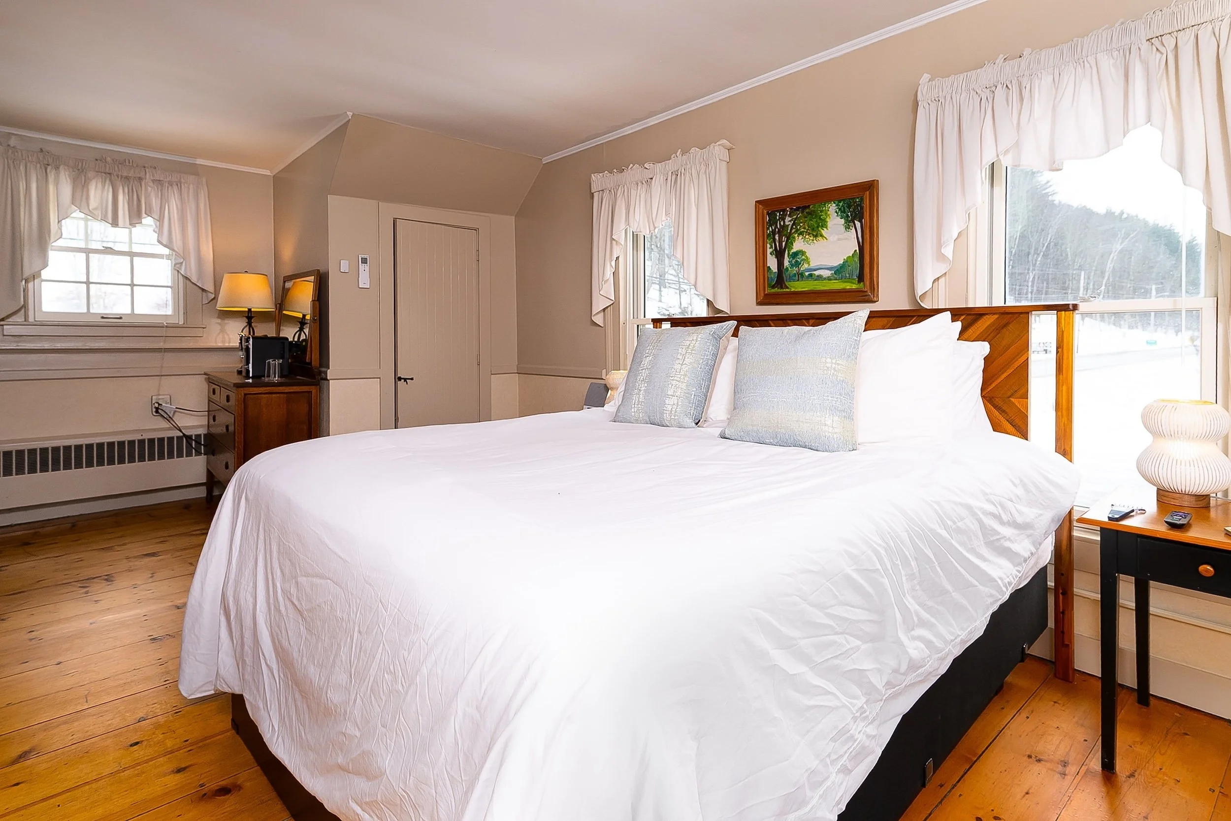 A bedroom with a wooden four-poster bed, red and white quilt, two bedside lamps, and seating area with two chairs, a small table, and a window showing a scenic view.