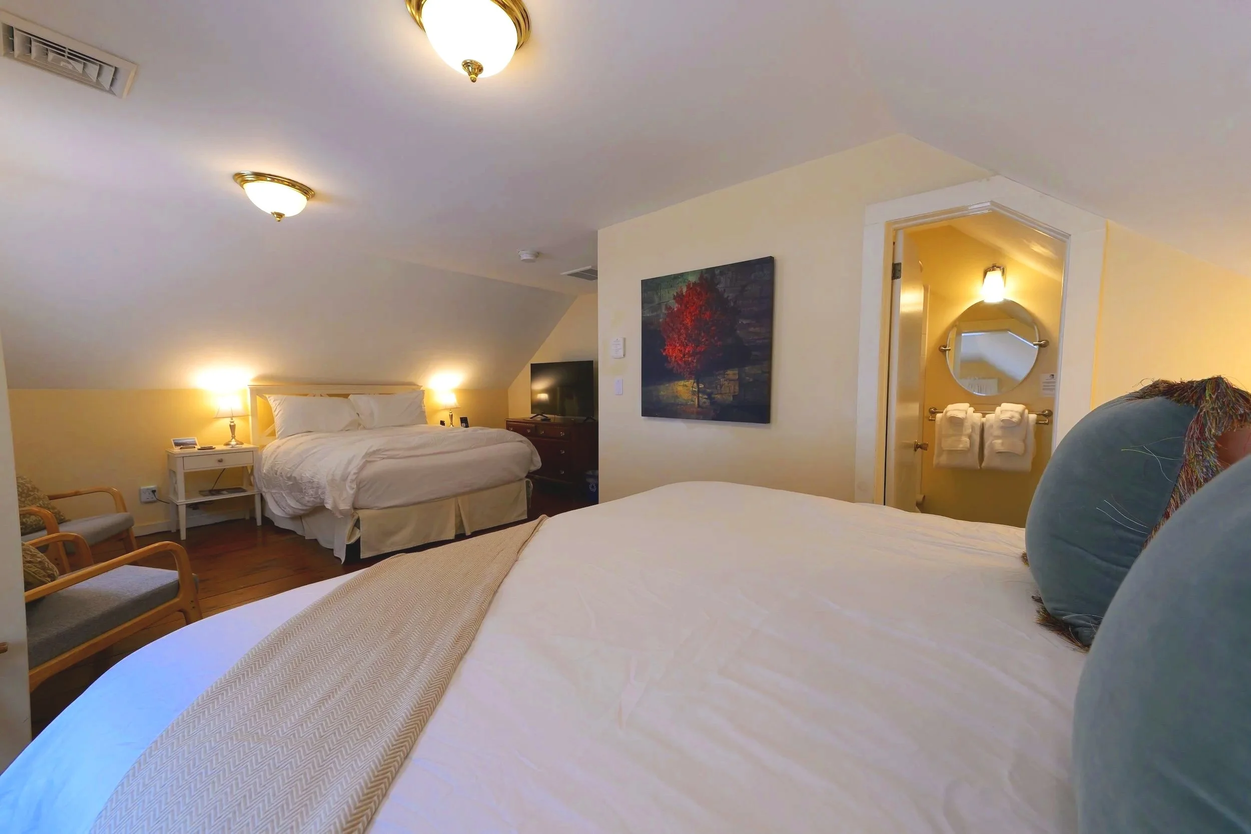 Two beds with white and yellow bedding, wooden floors, two windows with white curtains, two armchairs, lamps, a floral rug, and a small table with a coffee maker and glasses in a cozy hotel room.