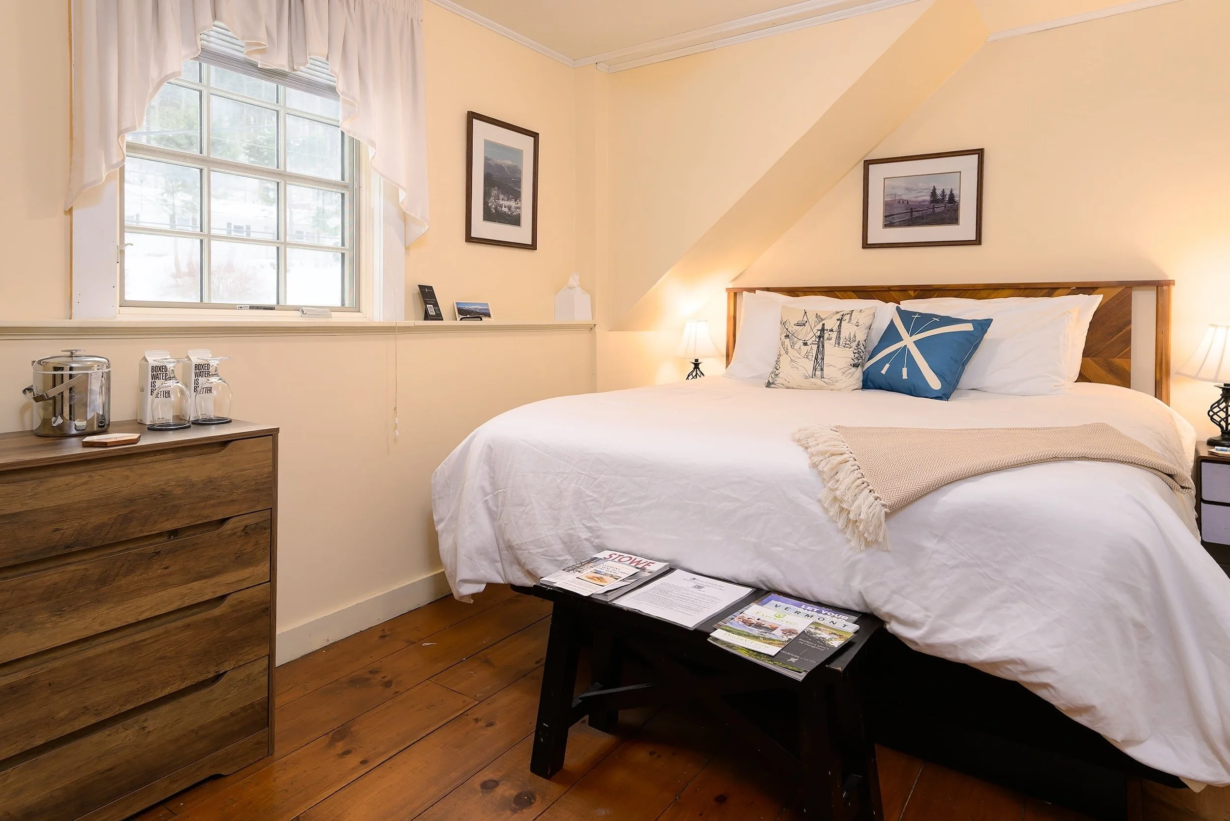 Cozy bedroom with wooden furniture, a bed with geometric patterned bedding, two lamps, and framed pictures on yellow walls. There is a window with white curtains and a wooden dresser with a mirror, coffee maker, and glasses.