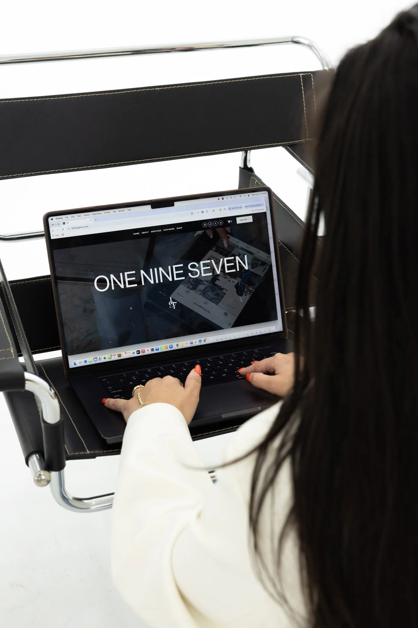 A woman with long dark hair and a white blazer sitting in a wheelchair, using a laptop with a website displaying the numbers 197 and 97.