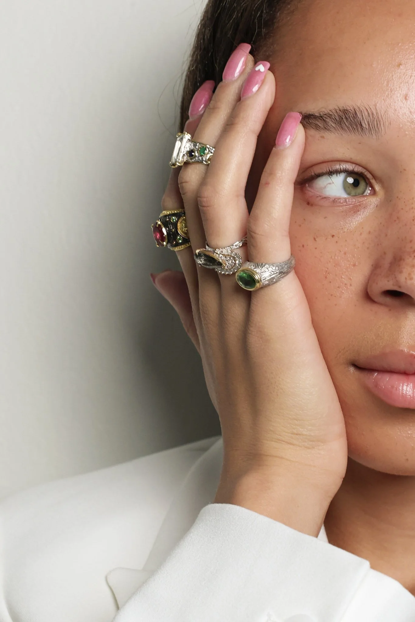 Close-up of a woman with green eyes and freckles, touching her face with her left hand. She has multiple jewelry rings on her fingers and pink nail polish with a small white heart design.