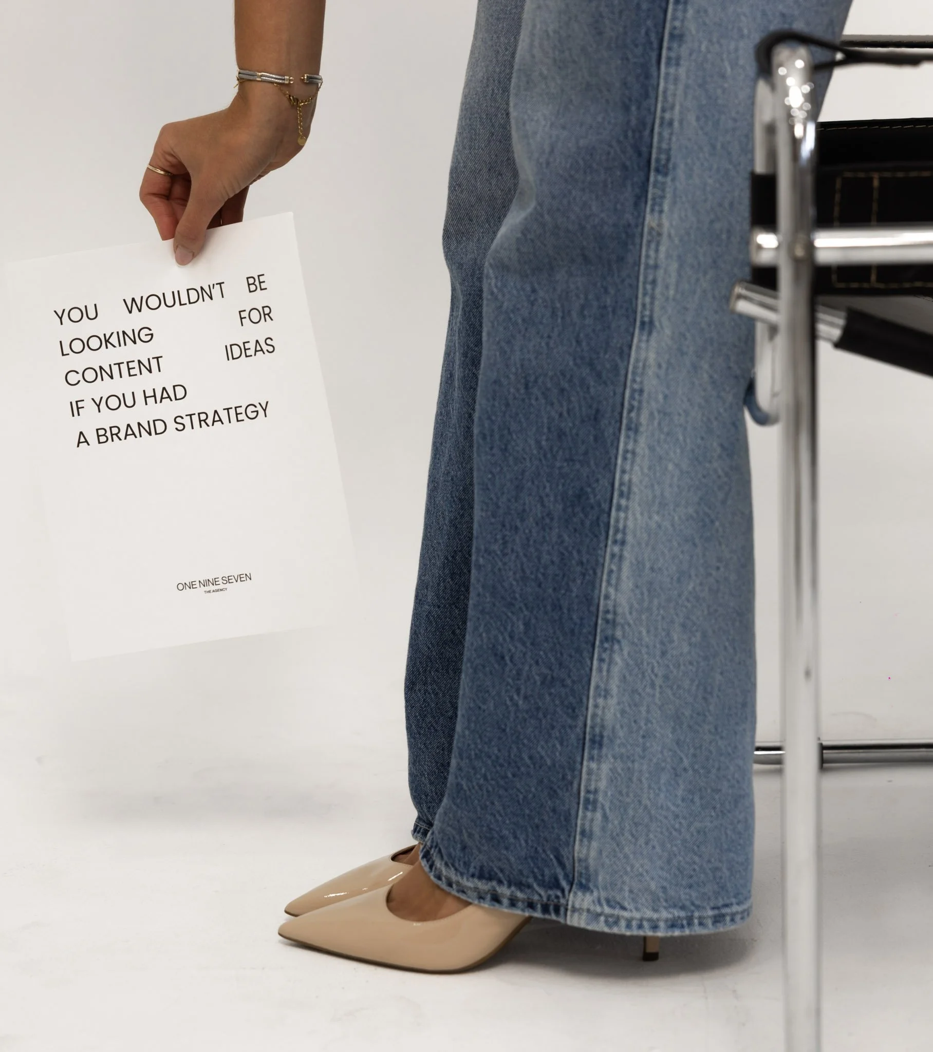 Close-up of a person standing, wearing blue jeans and beige high-heeled shoes, holding a white paper with black text, and a black shopping cart visible on the right side.