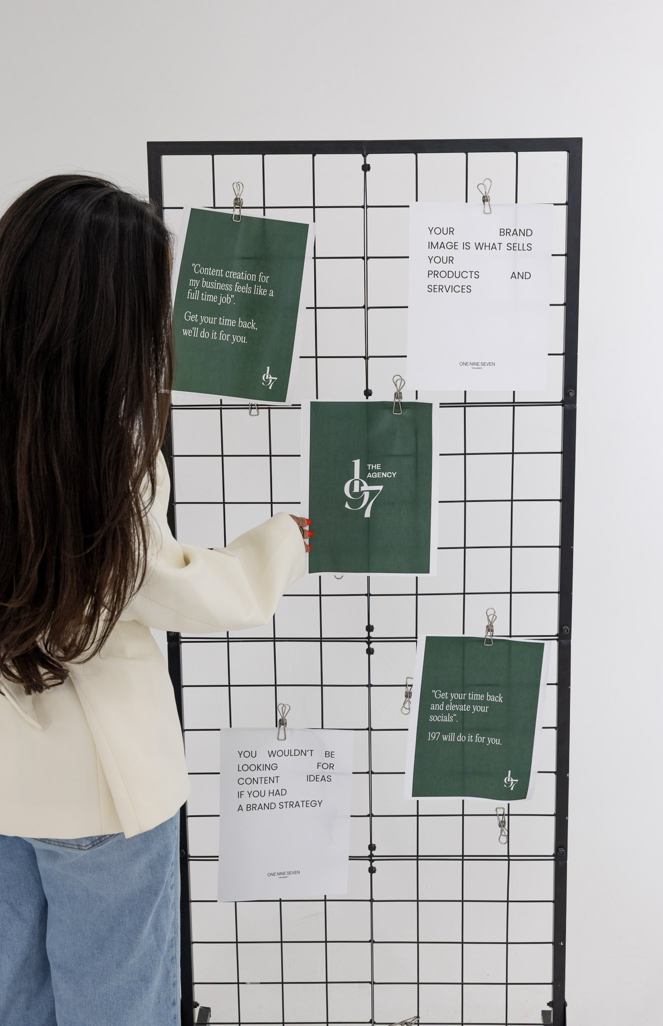 A person with long dark hair wearing a cream-colored blazer and blue jeans is arranging green and white paper notes on a black grid display. The notes contain texts related to branding and content creation, including quotes and statements about brand strategy and social media.