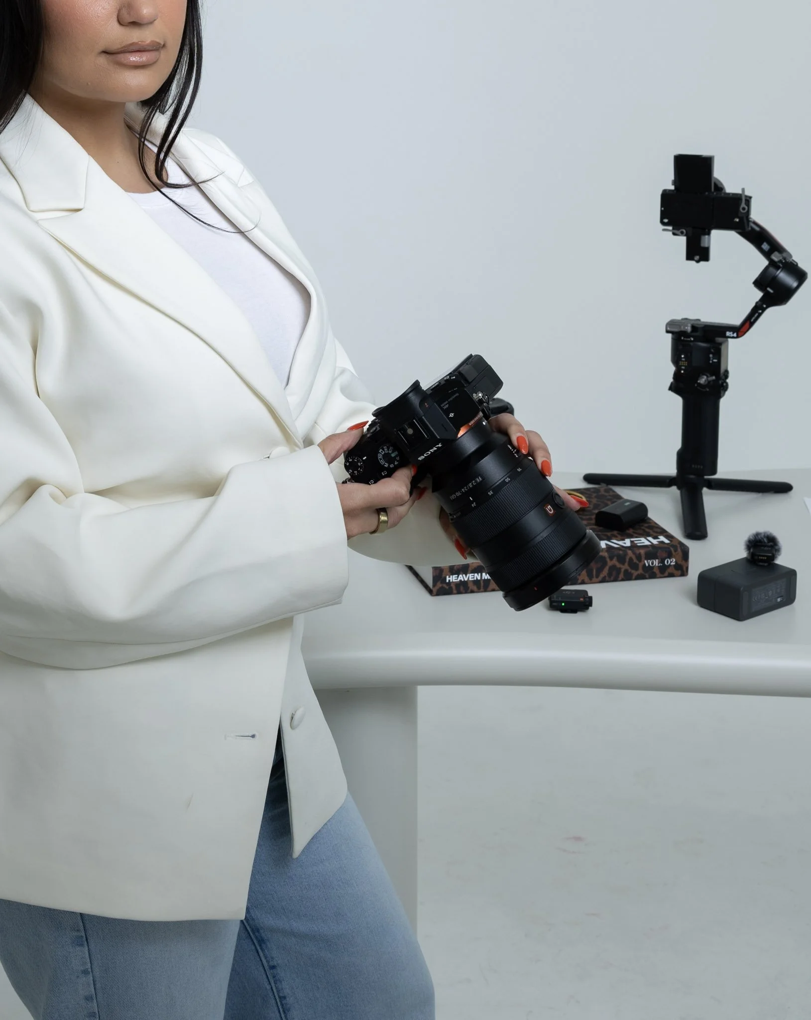 A woman wearing a white blazer and blue jeans, holding a camera, standing in front of a table with various camera equipment including a tripod, a box labeled 'HEAVEN' and 'VOL. 02', and other accessories.