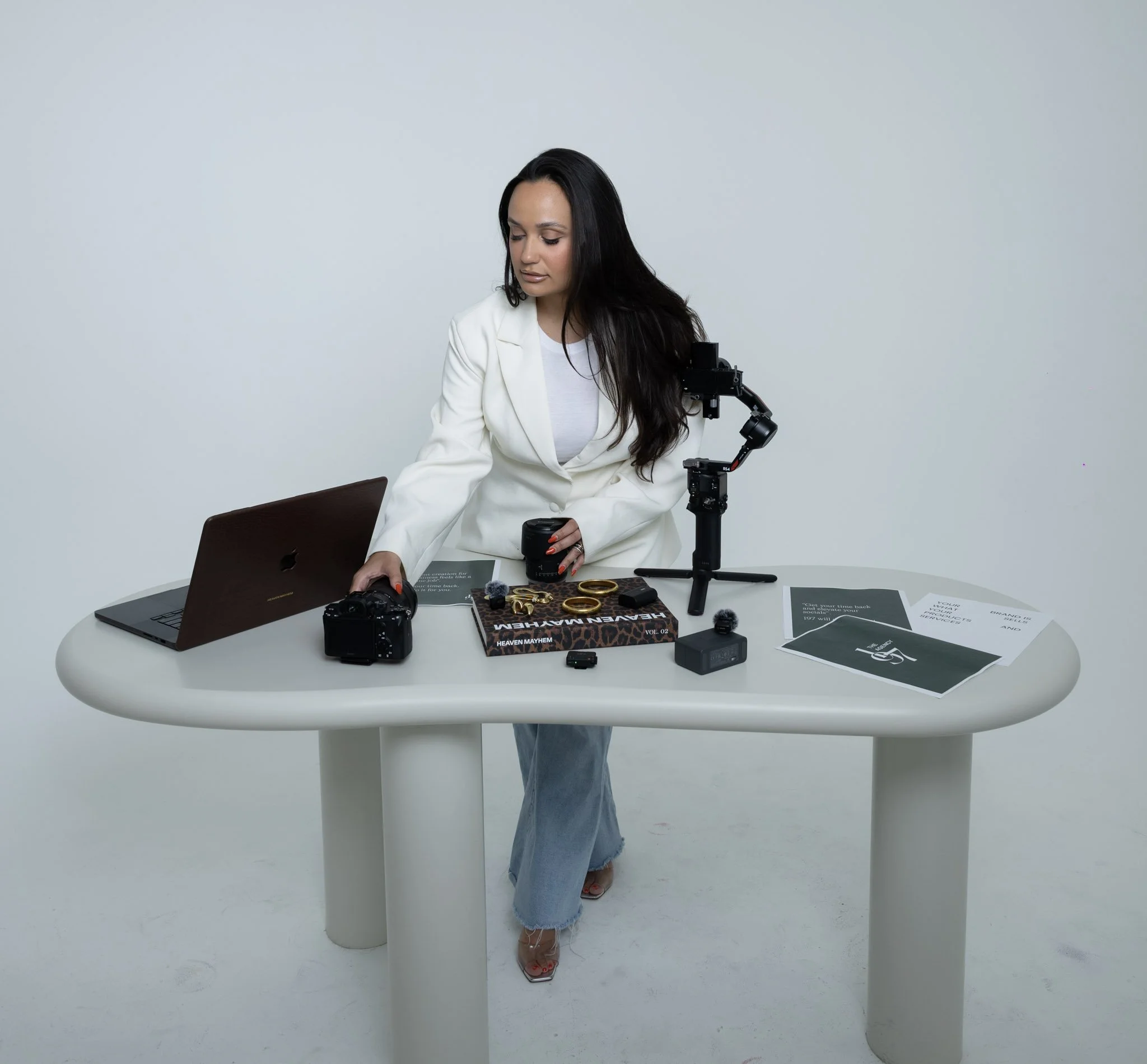 A woman with long dark hair wearing a white blazer and jeans is standing behind a white table with various electronic devices and magazines on it, in a minimalist setting with a plain gray background.
