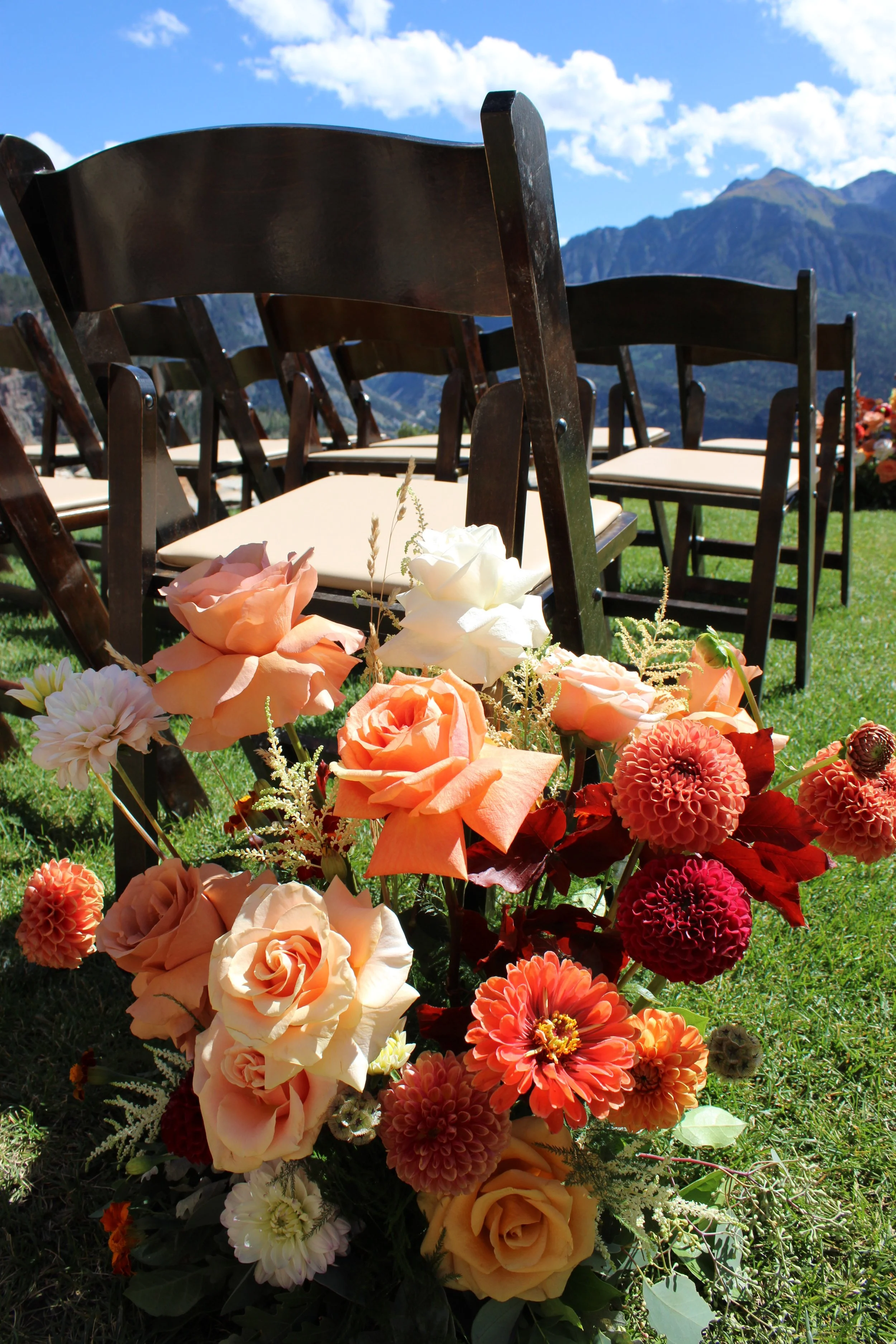 A floral arrangement with peach, white, and coral flowers in front of dark wooden chairs outdoors, with a mountain landscape and blue sky in the background.