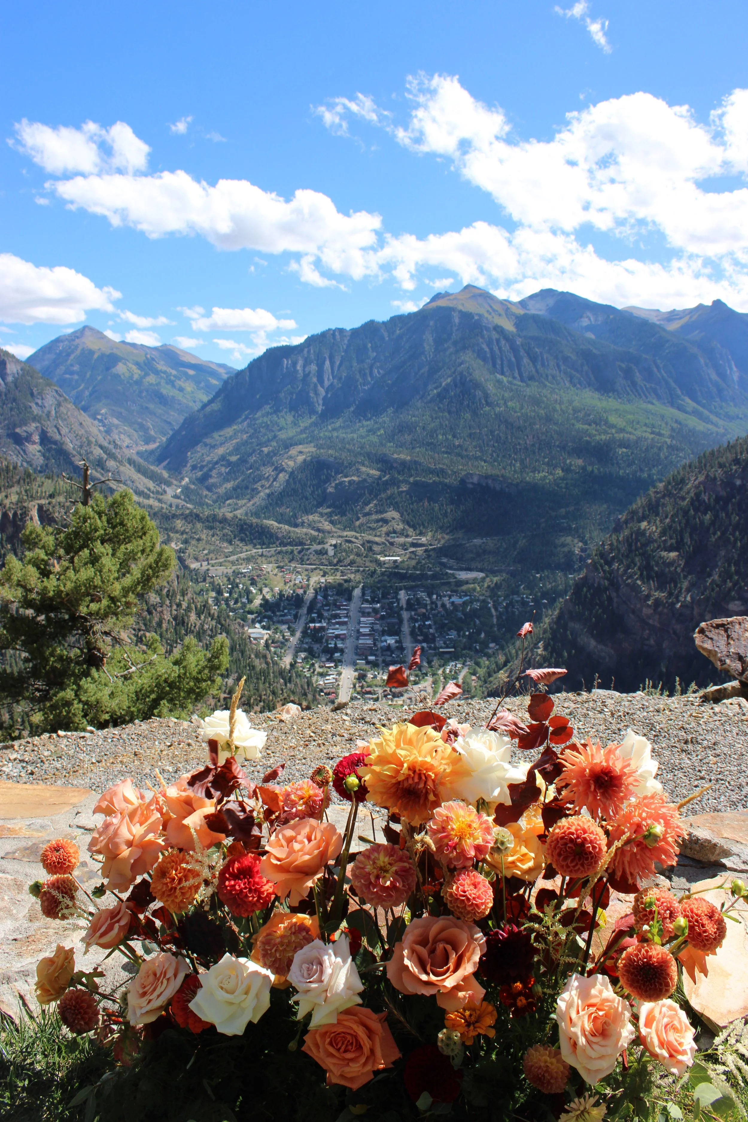 Colorful bouquet of flowers with white, peach, orange, and red blooms in the foreground on a stone surface. In the background, a scenic mountain landscape with a small town, lush green slopes, and a bright blue sky with scattered clouds.