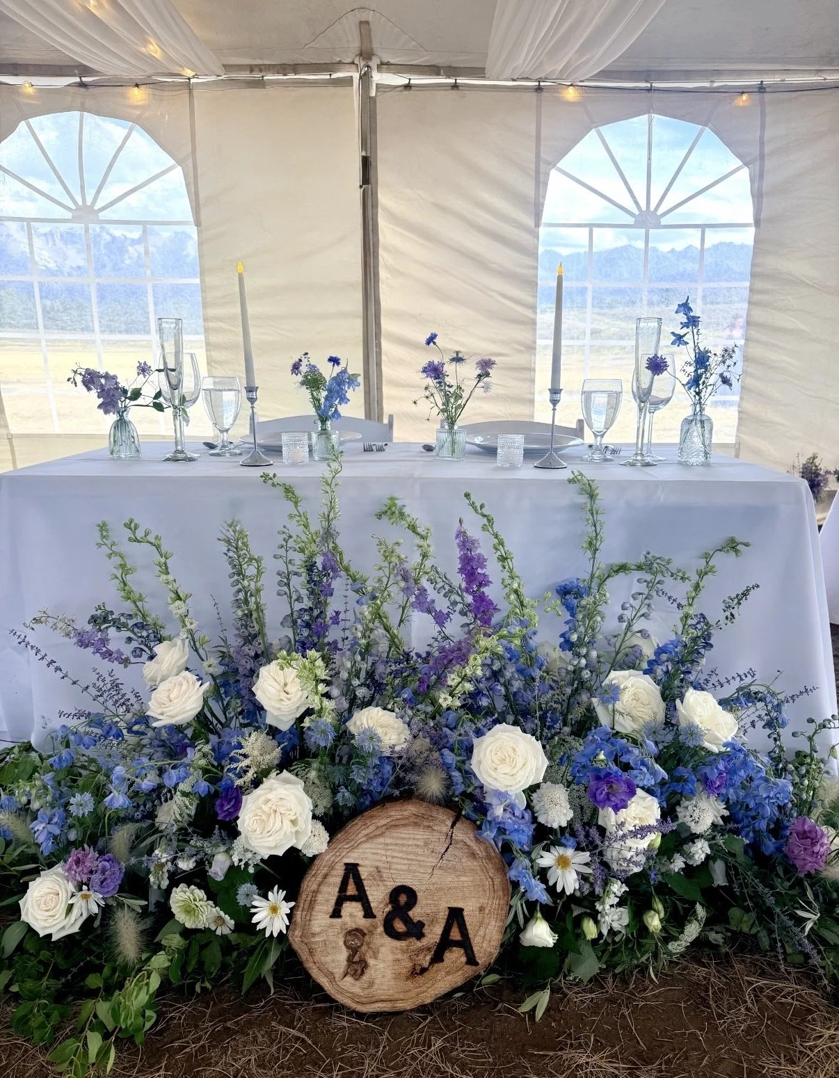 A decorated wedding or event table inside a tent with large window panels showing a mountain view. The table has a white tablecloth, floral centerpieces, tall candles, and glassware. In front of the table is a wooden log with the initials "A & A" eng
