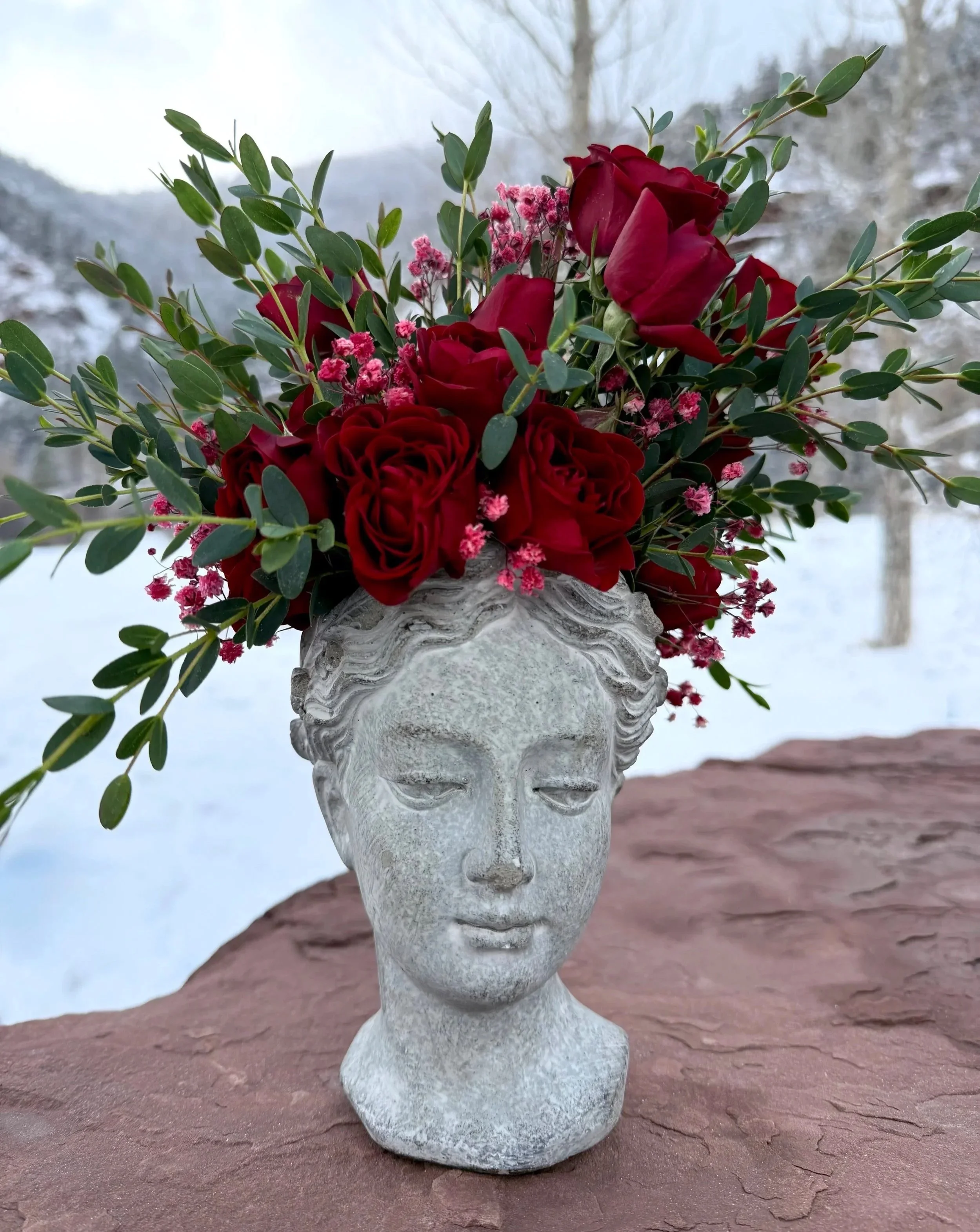 A bust-shaped stone vase filled with red roses, pink baby's breath, and greenery outdoors on a rock with a snowy background.