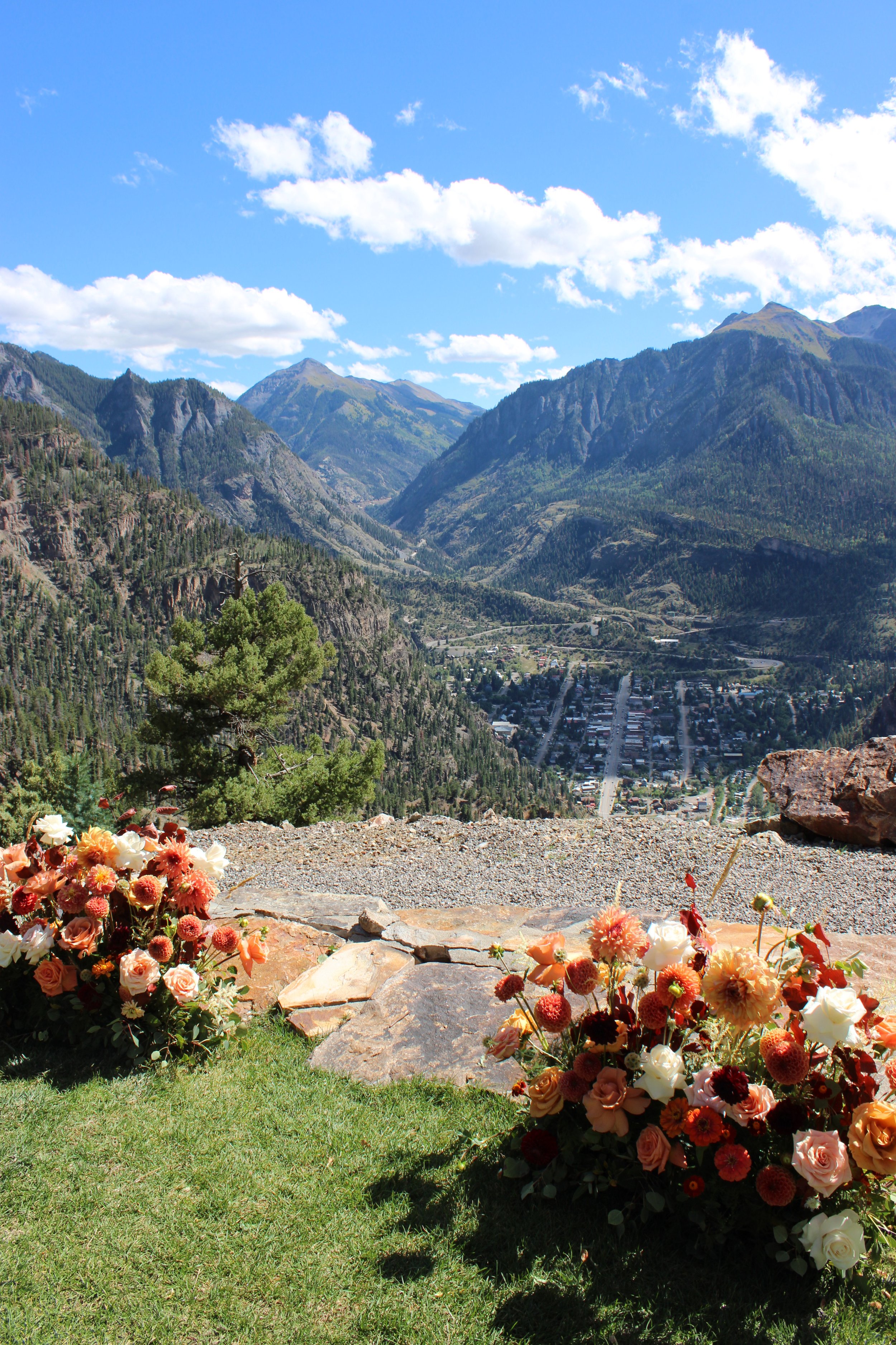 Colorful flower arrangement on rocks in the foreground with a mountain landscape and blue sky with clouds in the background.