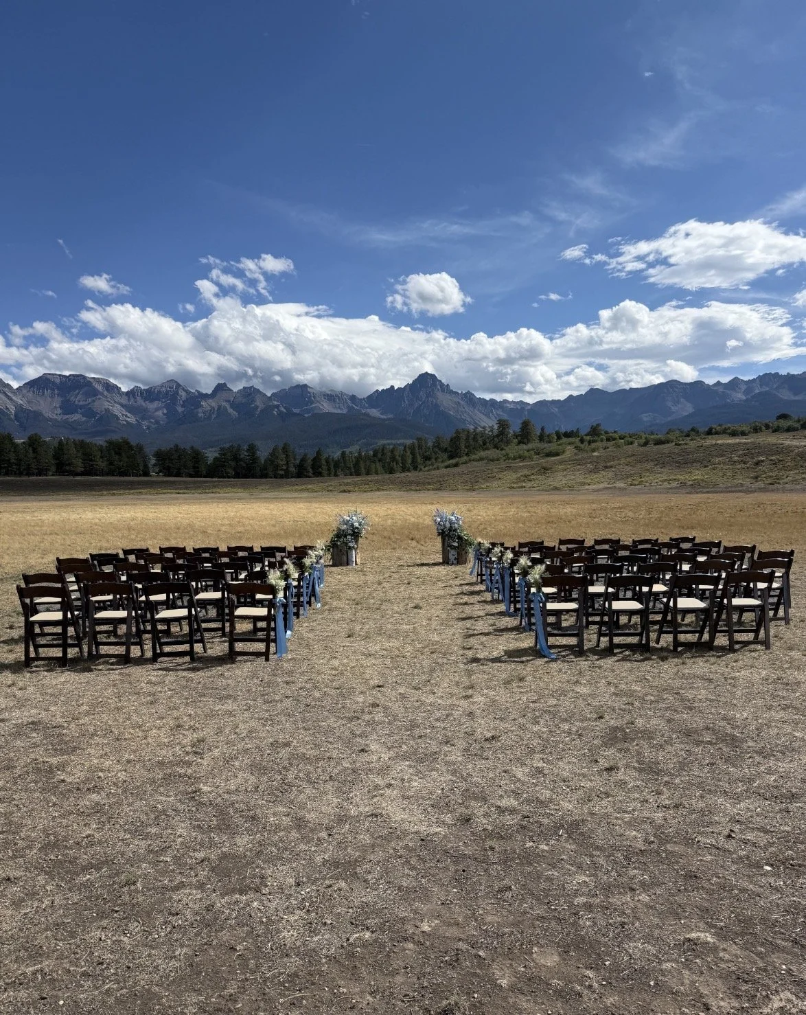 Outdoor wedding ceremony setup with chairs and floral decorations in a field with mountain range and blue sky in the background.