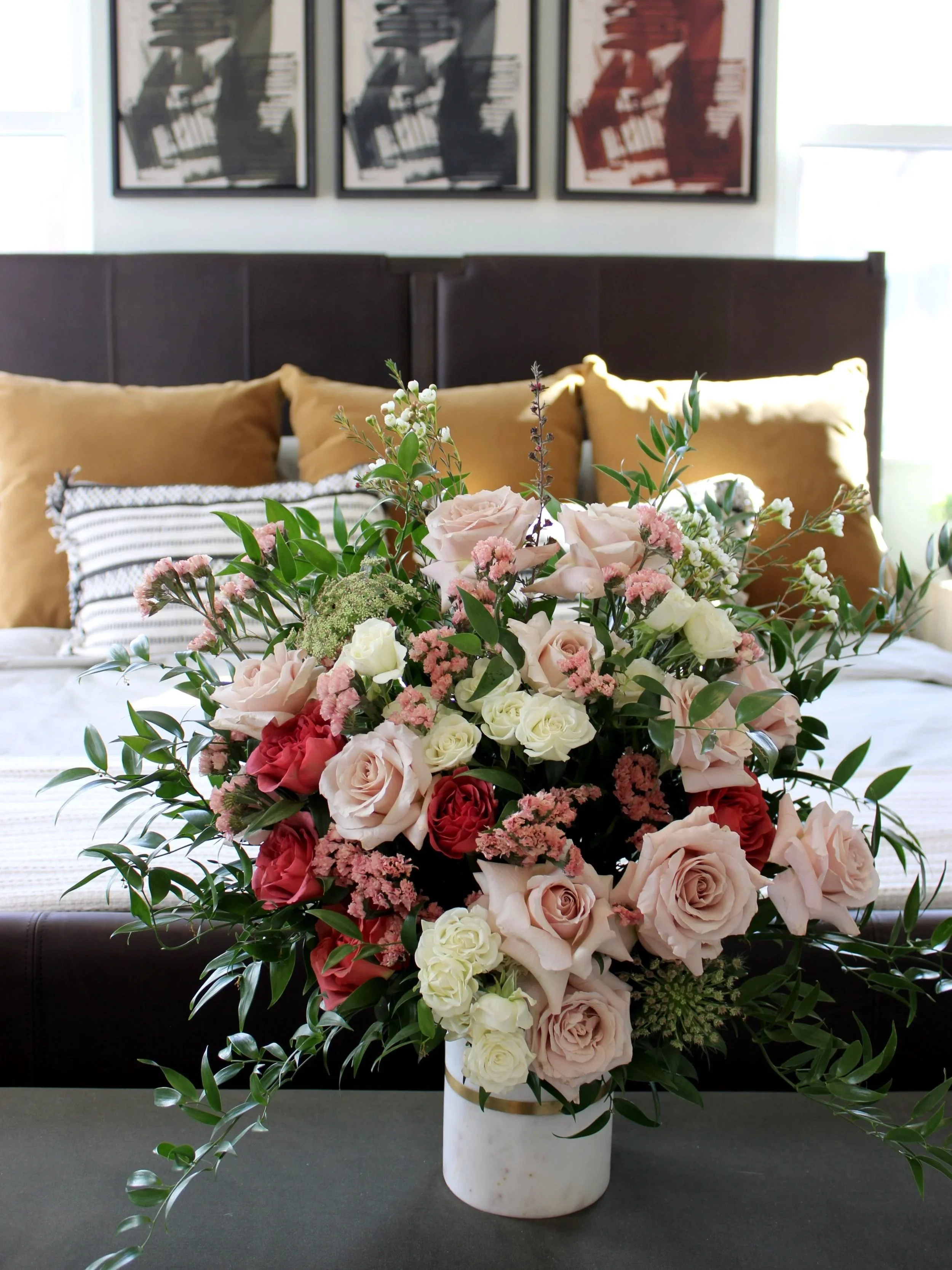 A large bouquet of pink, white, and red roses along with other greenery in a white vase on a dark table, with a bed and framed artwork in the background.