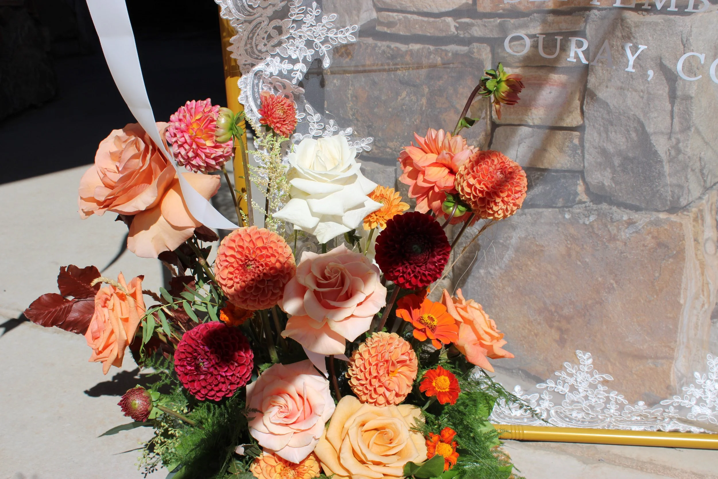 A floral arrangement with pink, orange, cream, and red flowers in front of a stone wall and a translucent sign.