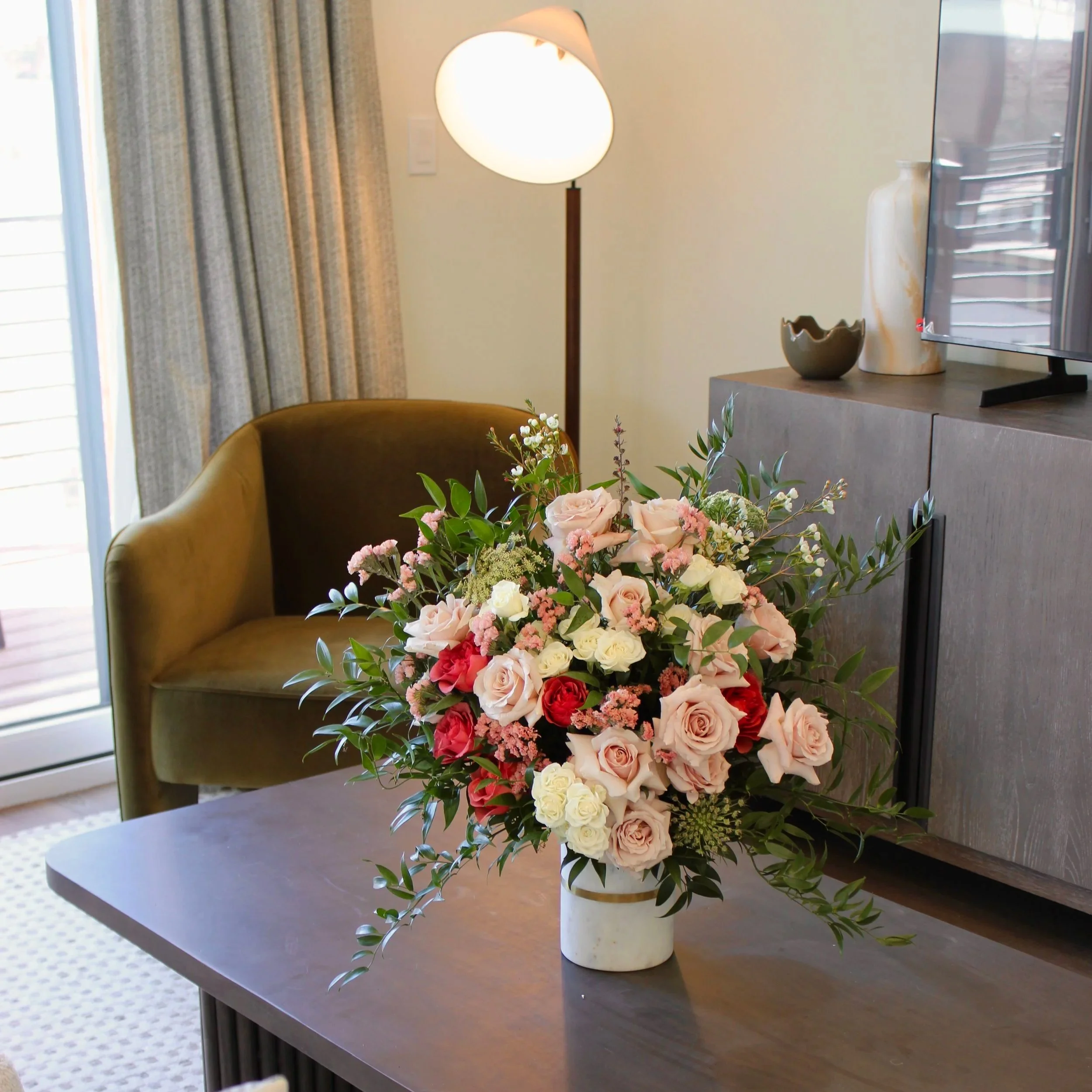 A cozy living room corner with a brown armchair, a floor lamp, and a modern wooden cabinet. A large bouquet of pink, red, and white roses with greenery is placed on a dark wood table in the foreground.
