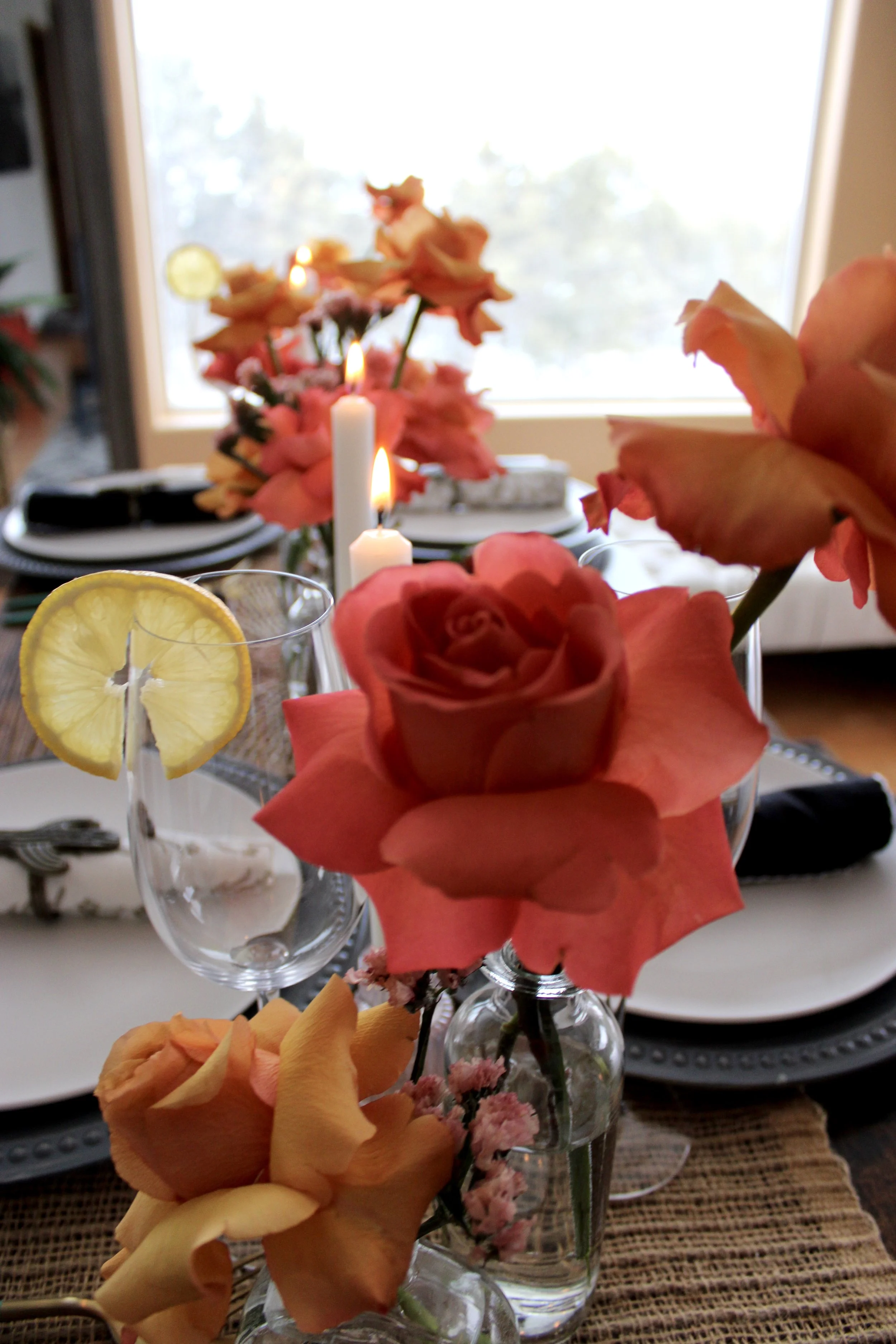 Dining table set with floral centerpiece, candles, water glasses with lemon slices, and gray plates and utensils, in front of a window with trees outside.