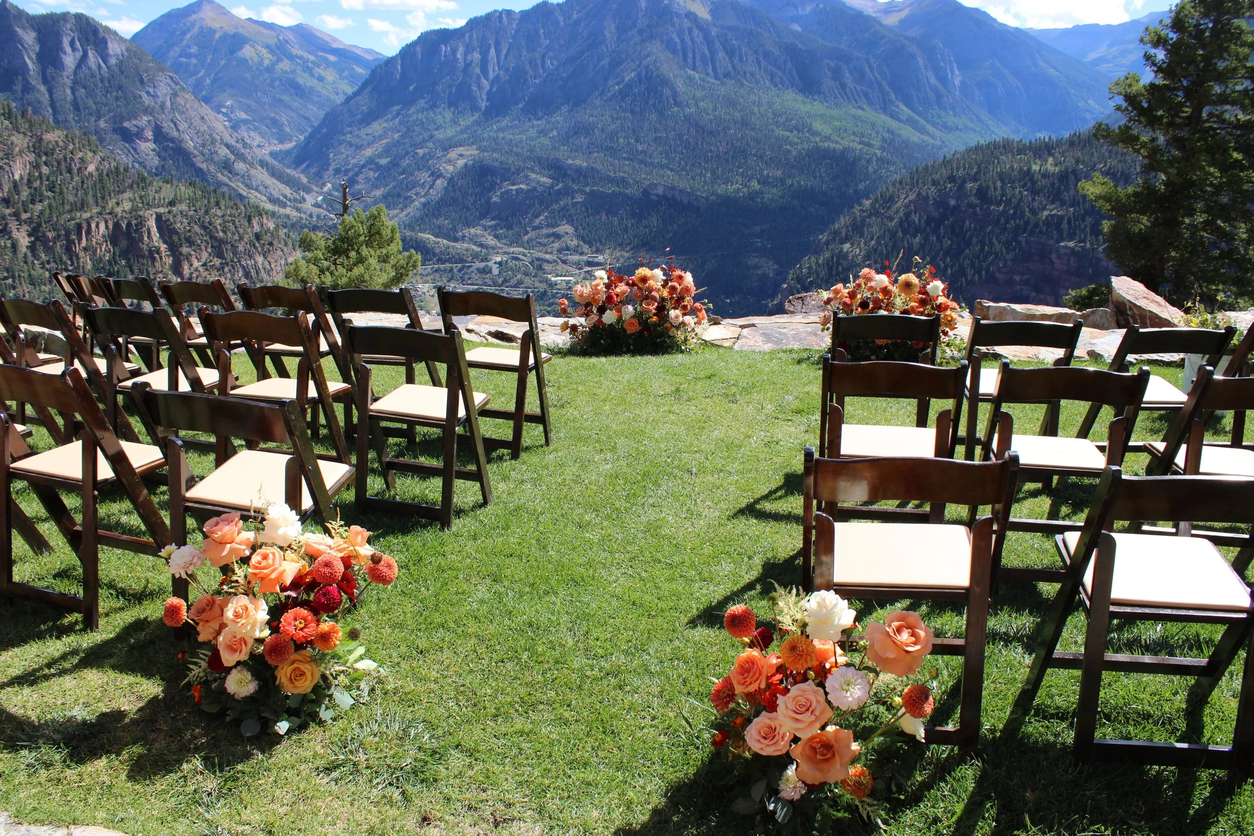 Arranged chairs and flower arrangements set up outdoors on a grassy area overlooking mountain scenery.