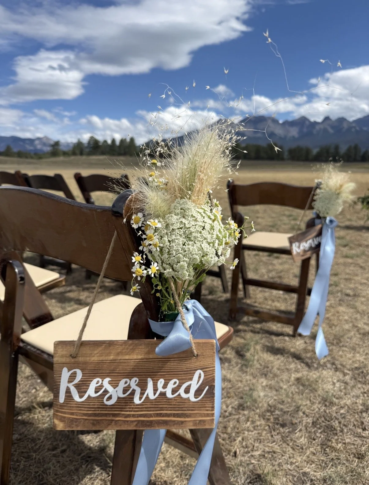 Wooden chairs with floral arrangements and a reserved sign at an outdoor event space with mountains in the background.
