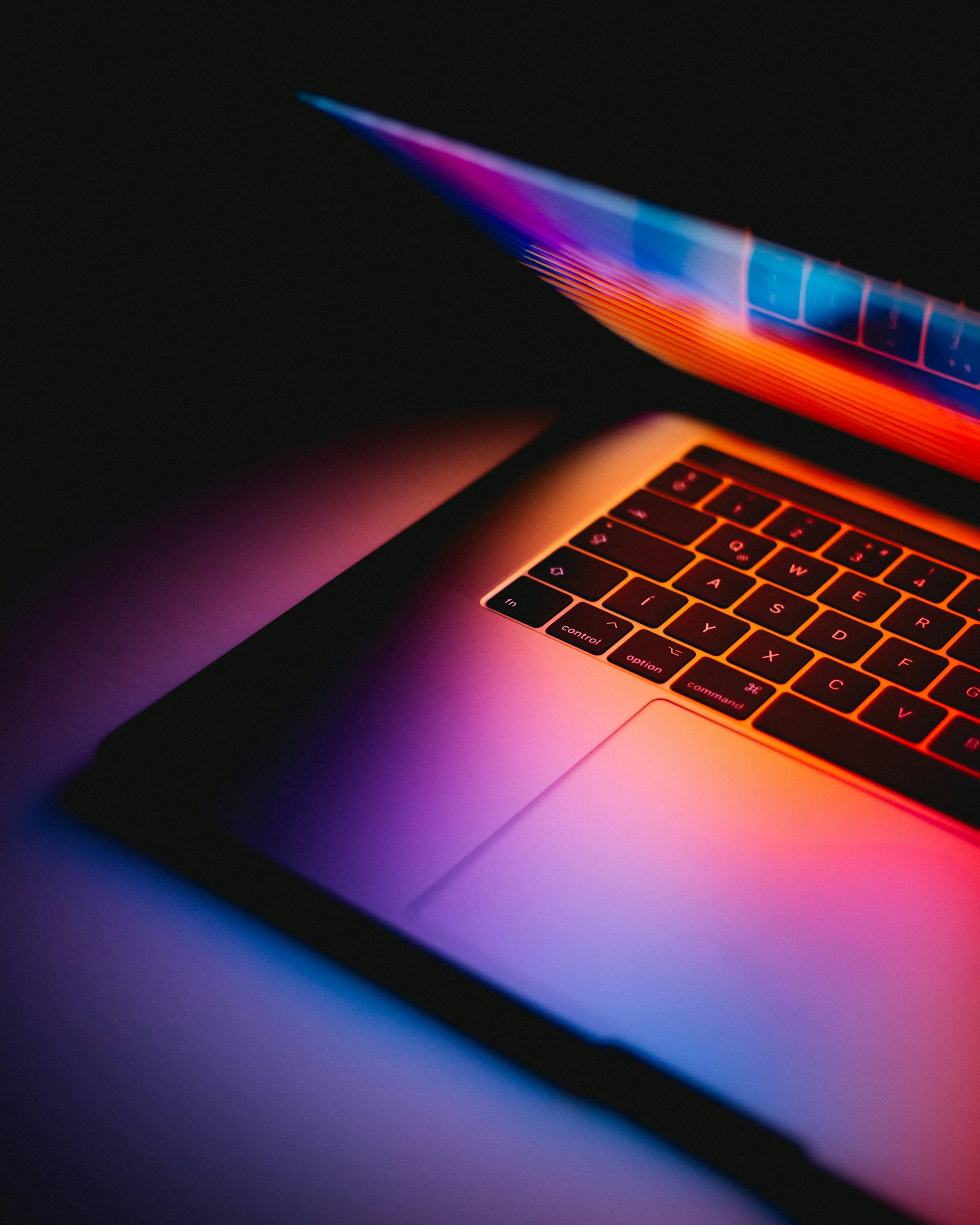 A close-up of a laptop keyboard and screen illuminated with vibrant red, orange, blue, and purple neon lights in a dark environment.