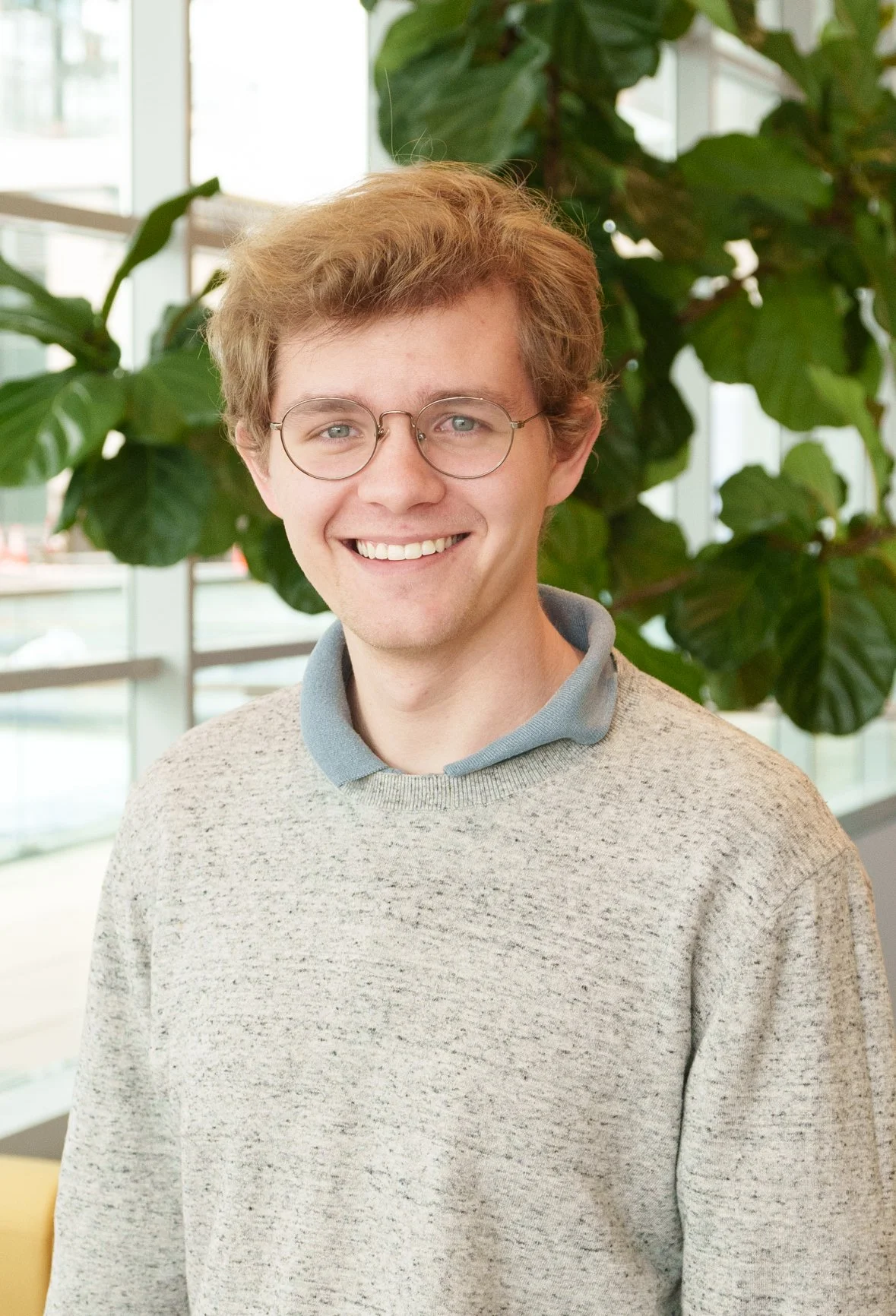 Person wearing glasses and a gray sweater, standing indoors with green plants in the background.