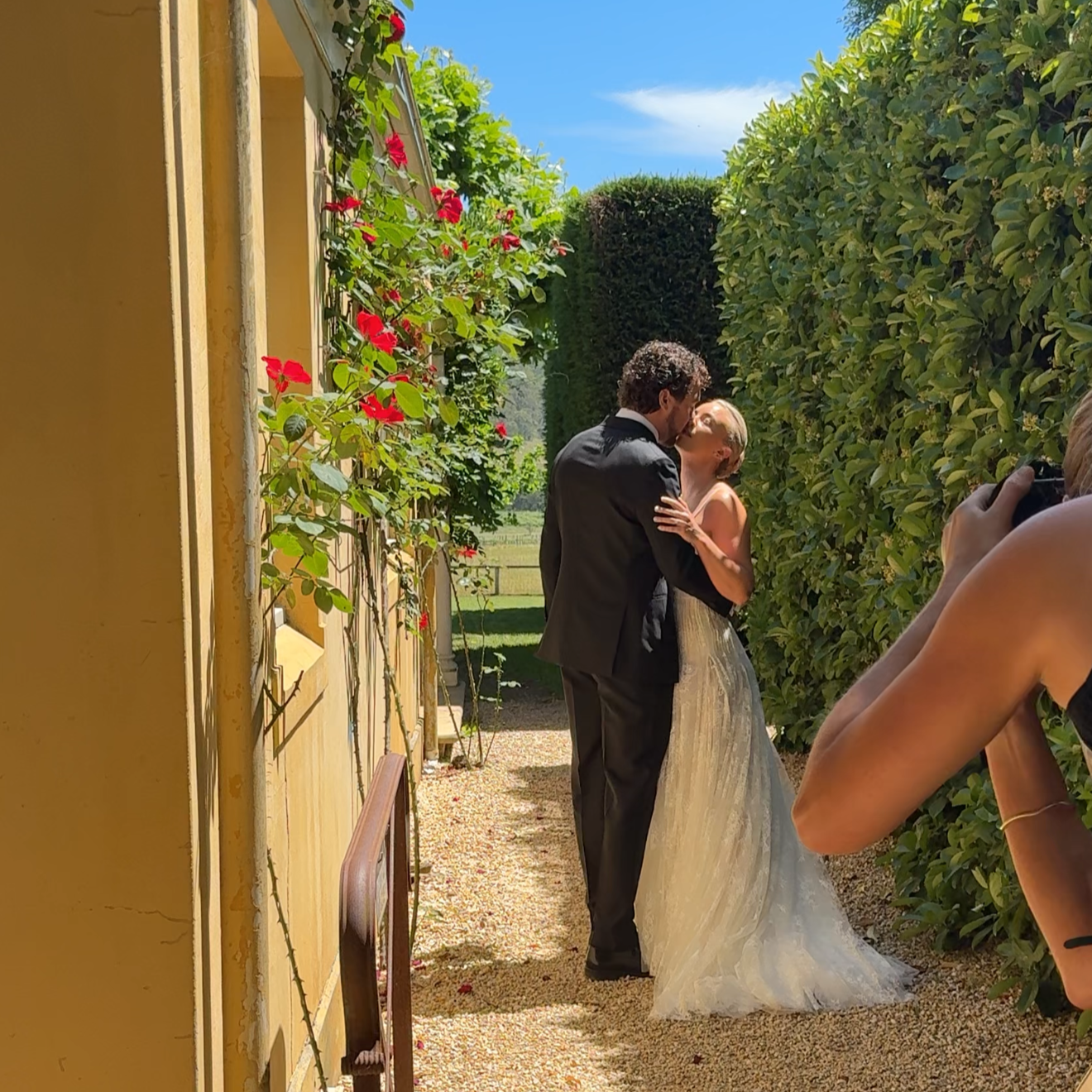 A bride and groom kiss outdoors on a sunny day, with a photographer capturing the moment. The couple is in a garden pathway bordered by greenery and flowers.