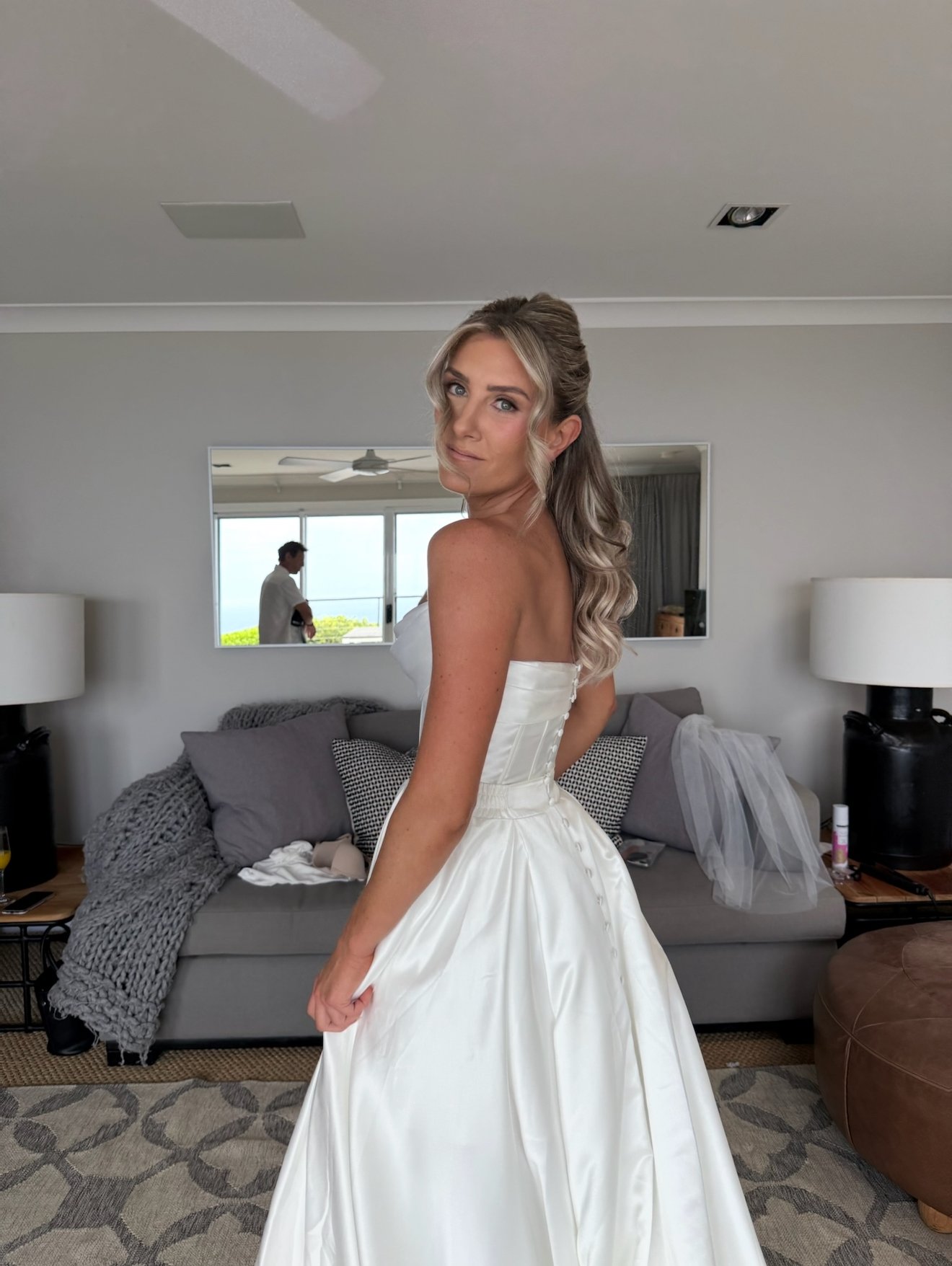 A woman in a white wedding dress standing in a living room, with a mirror behind her reflecting a man near a window.
