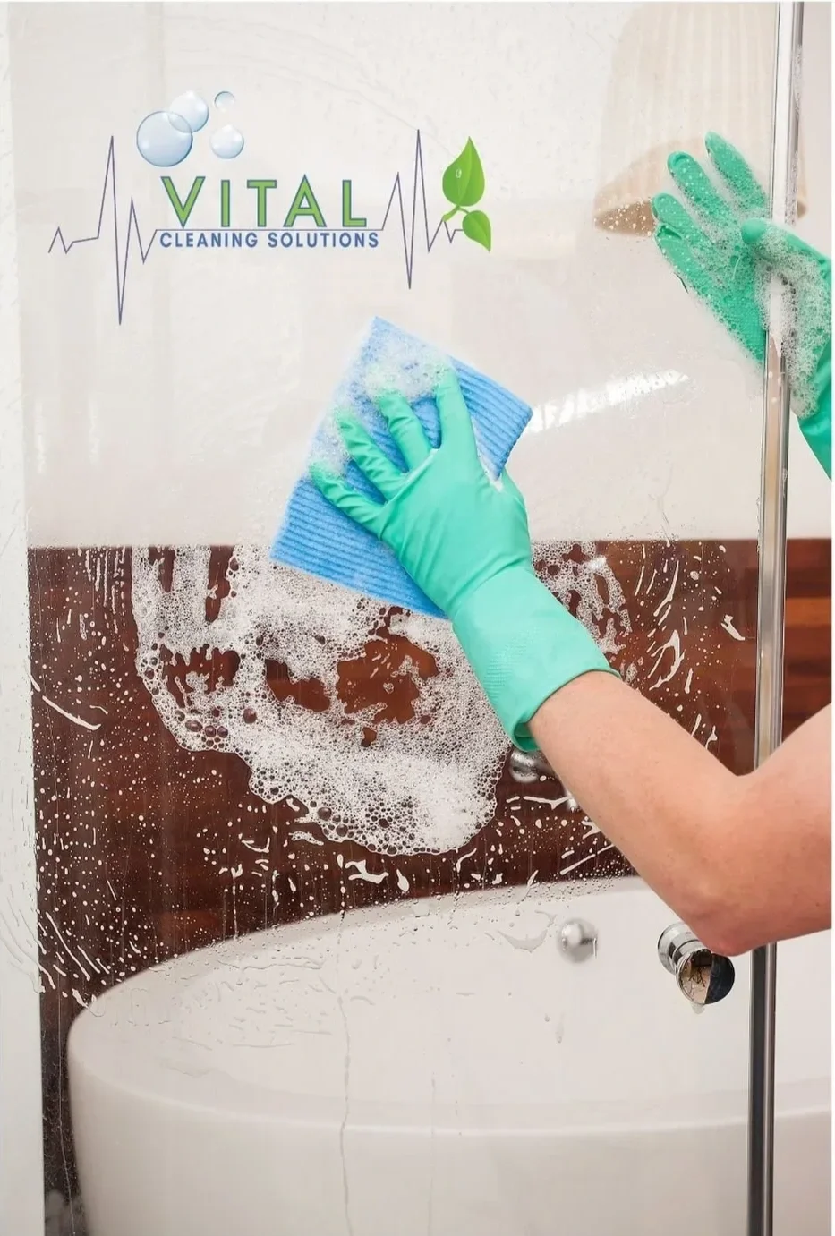 A person cleaning a glass shower door with a blue sponge and wearing green gloves, with foam and soap suds on the glass, in a bathroom with a white bathtub and brown wall panel.