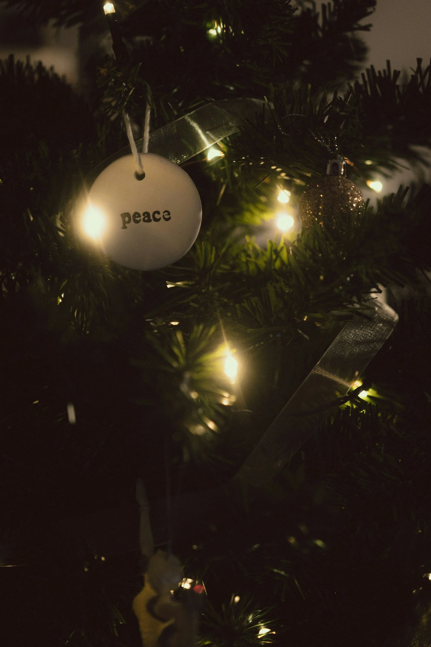 A Christmas tree decorated with lights and ornaments, including a round white ornament with the word 'peace' written on it and a glittery gold ball ornament.