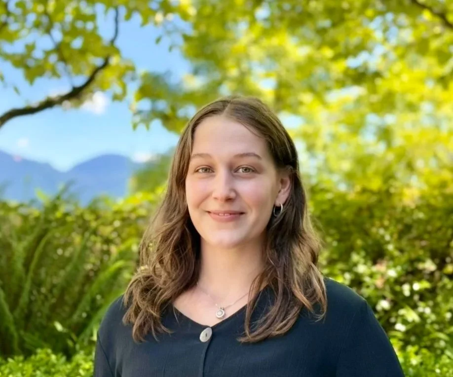 Young woman with shoulder-length brown hair smiling outdoors under green trees with mountains in the background.