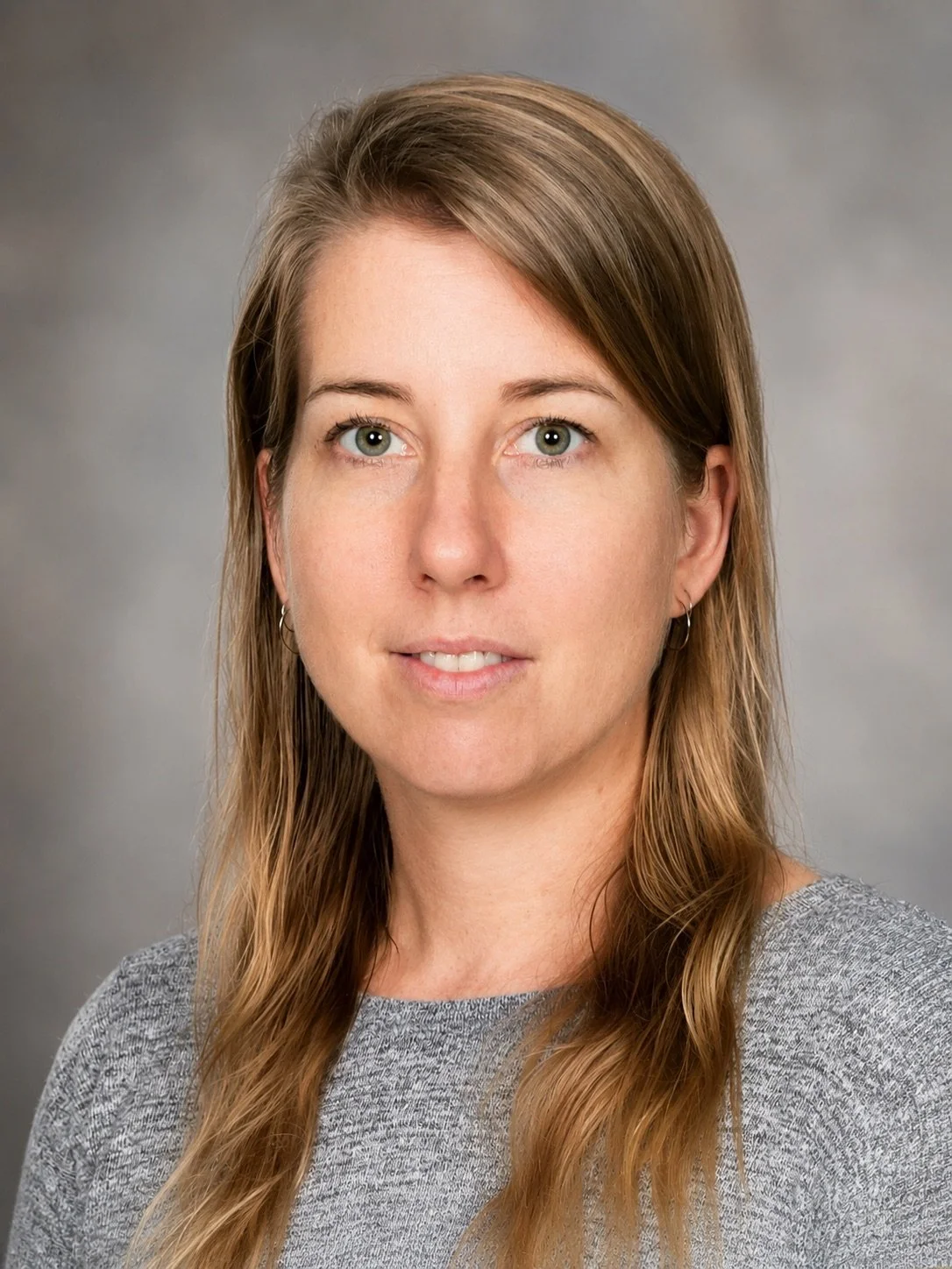 Headshot of a woman with long wavy light brown hair, green eyes, and fair skin, wearing a gray top and small hoop earrings, against a gray background.