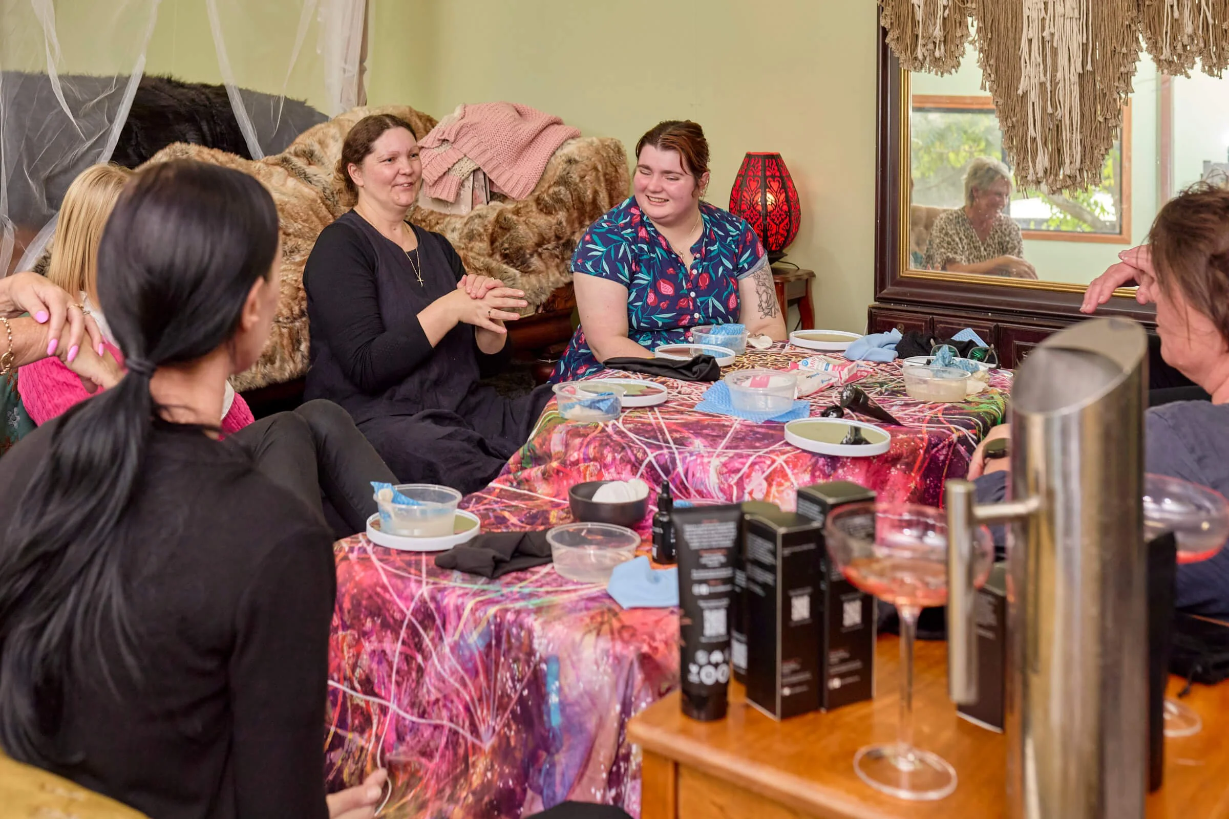 Group of women sitting around a table, sharing a conversation, with makeup supplies and drinks on the table.
