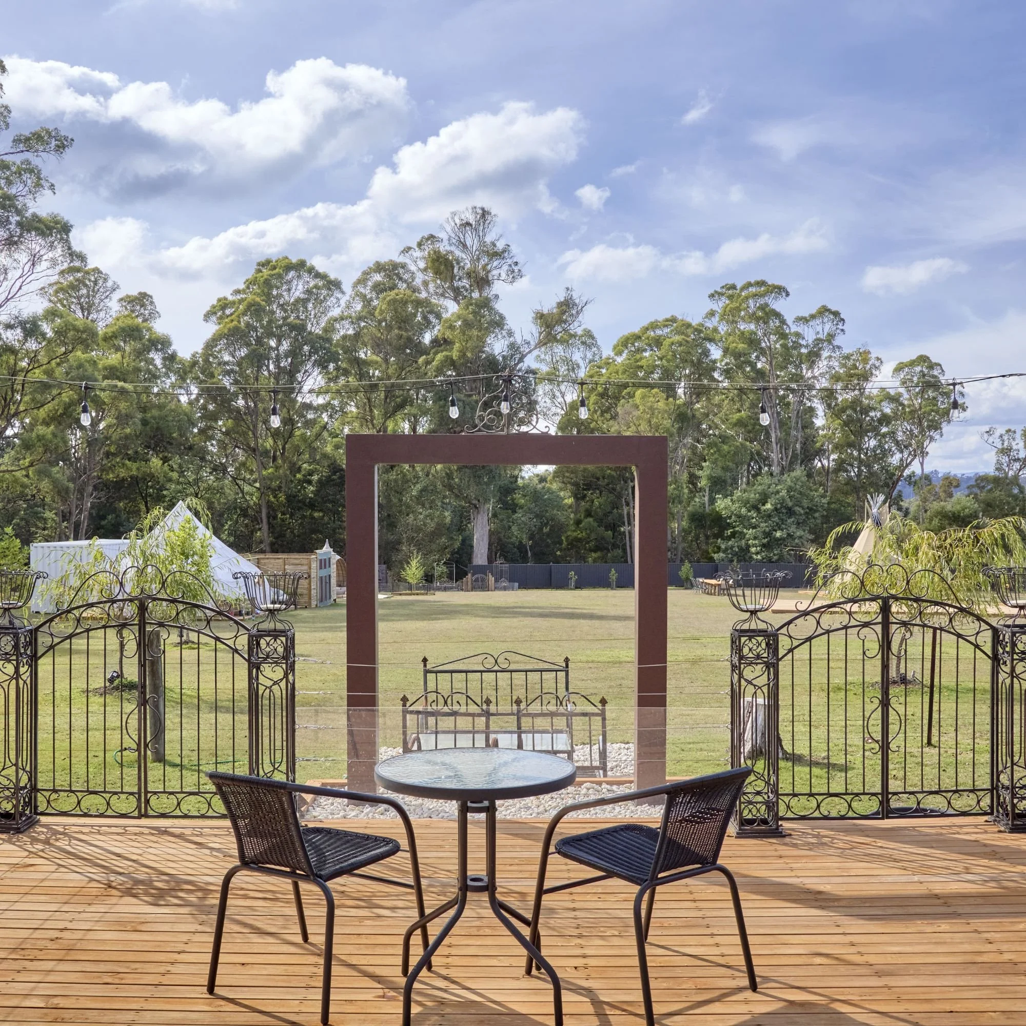 Outdoor patio with black metal table and two chairs, enclosed by a decorative metal fence, overlooking a grassy yard with trees in the background, string lights hanging overhead, under a partly cloudy sky.
