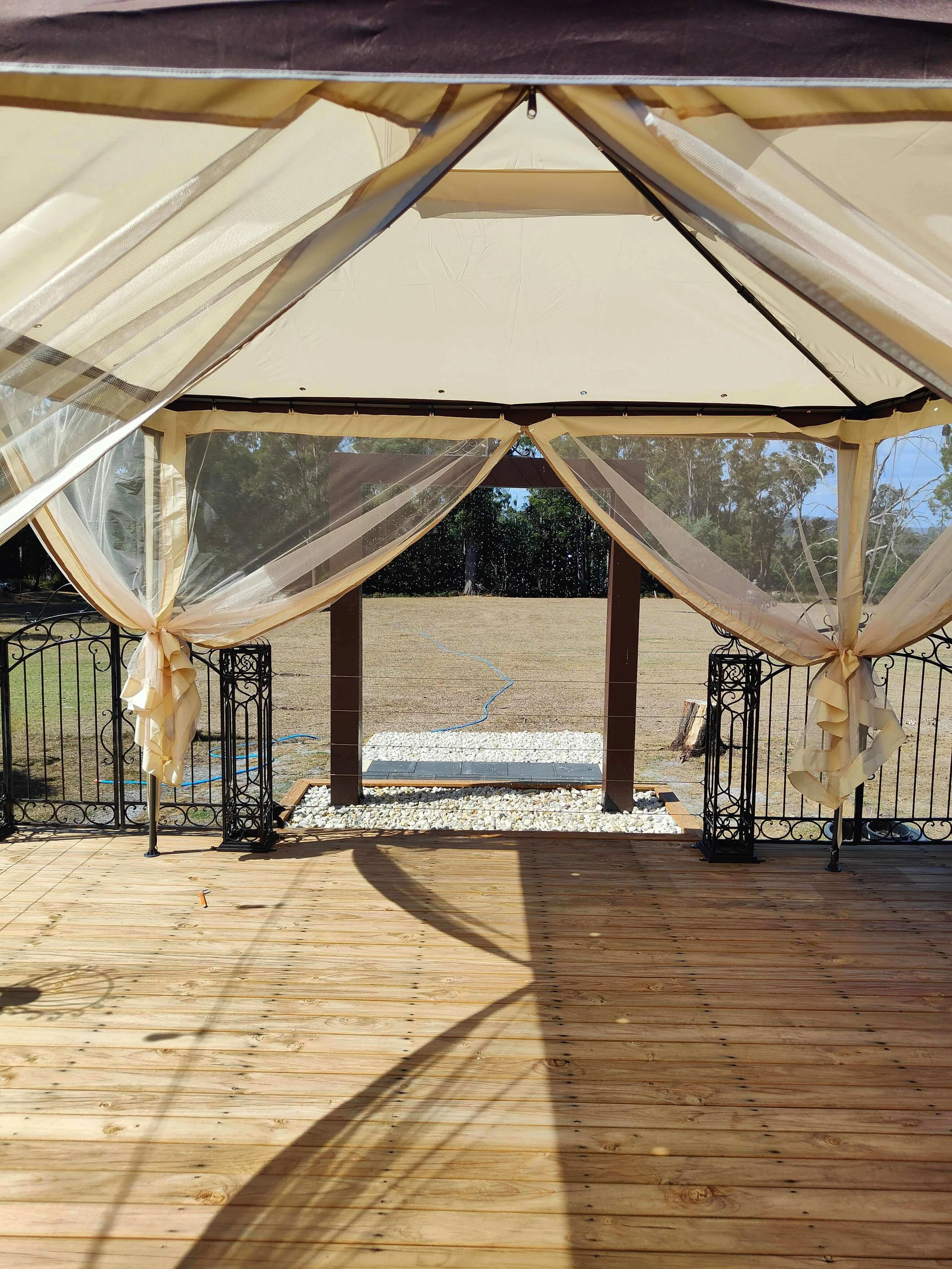 A screened outdoor gazebo with beige curtains tied to the sides, overlooking a grassy yard with a small stone pathway, trees in the background, and a wooden deck in the foreground.