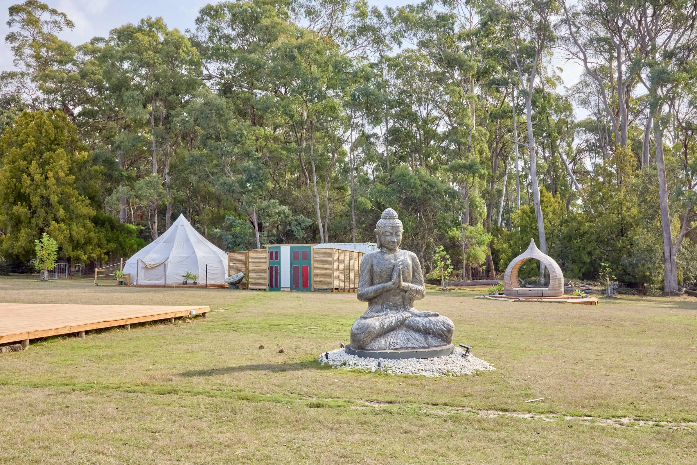 Outdoor scene with a large seated Buddha statue in the foreground, a white tent, small structures, and trees in the background.