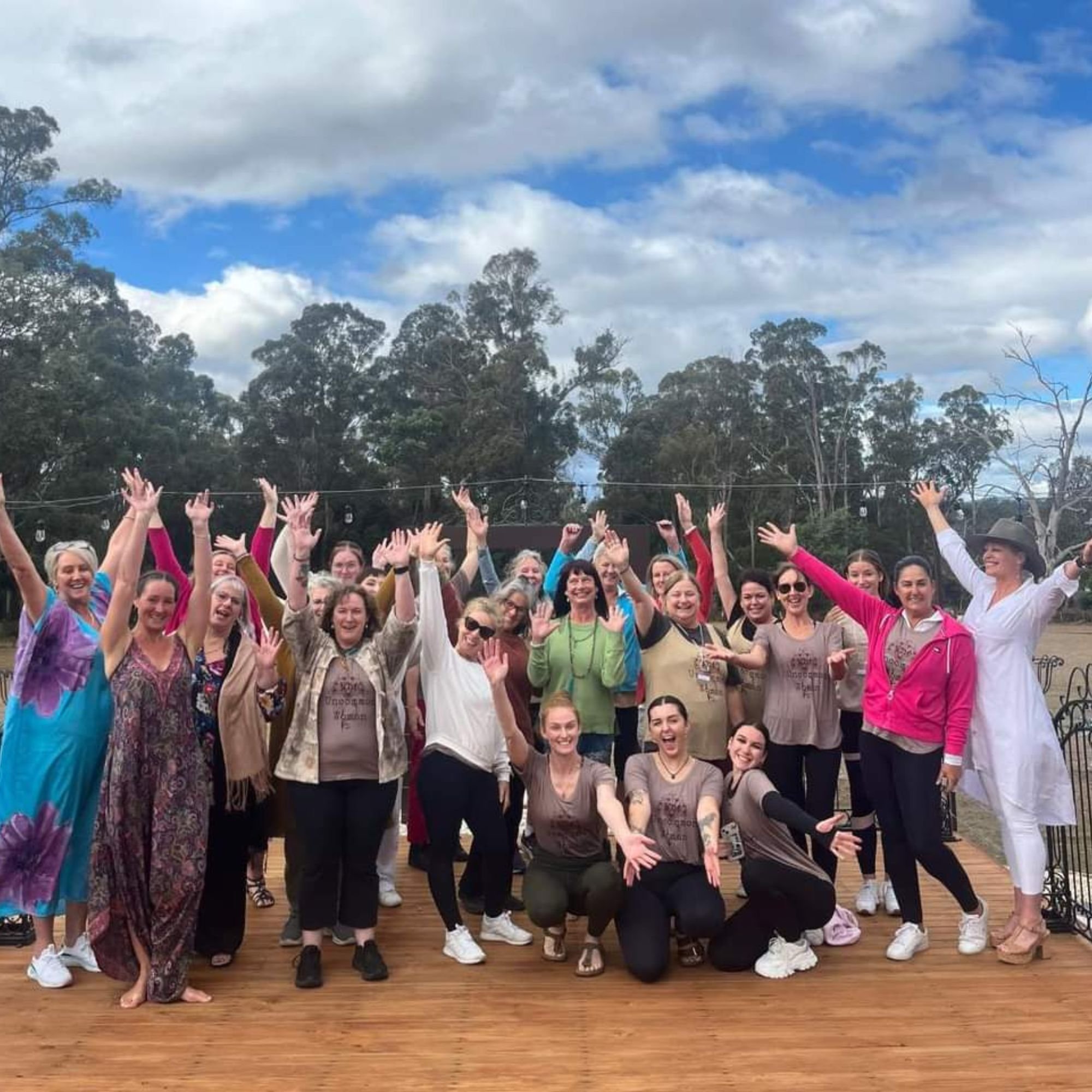 Group of women outdoors on a wooden deck, raising their hands and smiling, against a background of trees and cloudy sky.
