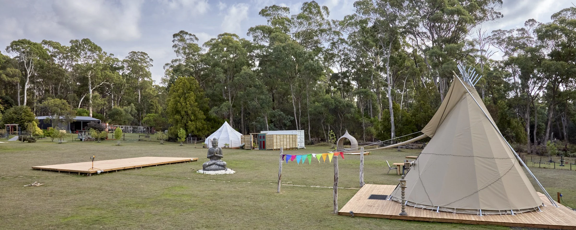 Outdoor scene with a teepee tent and a Buddha statue in a grassy area, surrounded by trees and various small structures, with colorful buntings hanging between wooden posts.