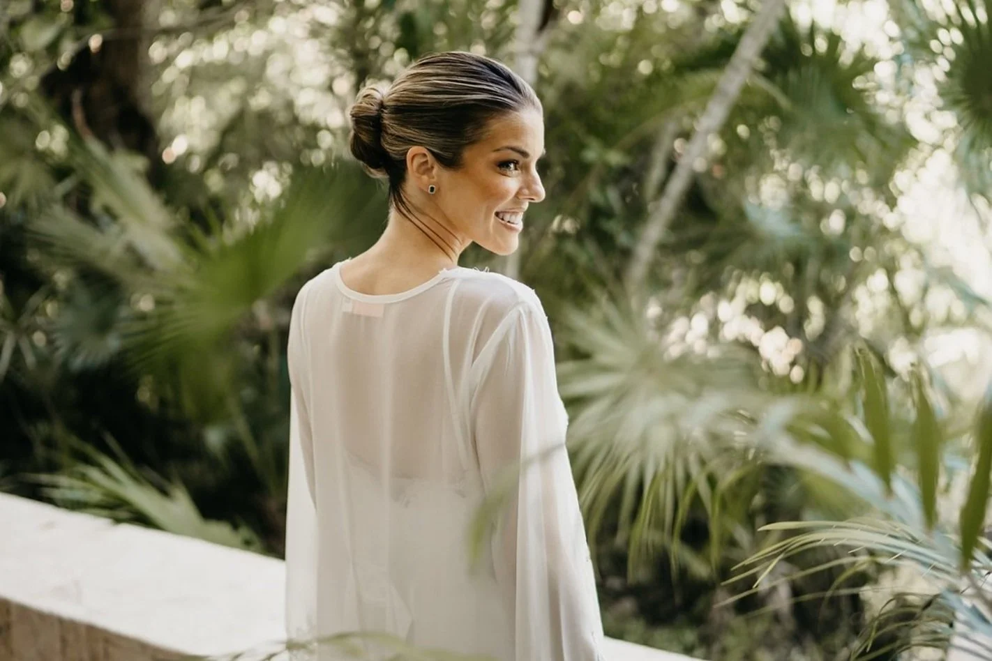 A woman with dark brown hair tied in a bun, smiling and looking over her shoulder outdoors among green tropical plants, wearing a sheer white blouse.