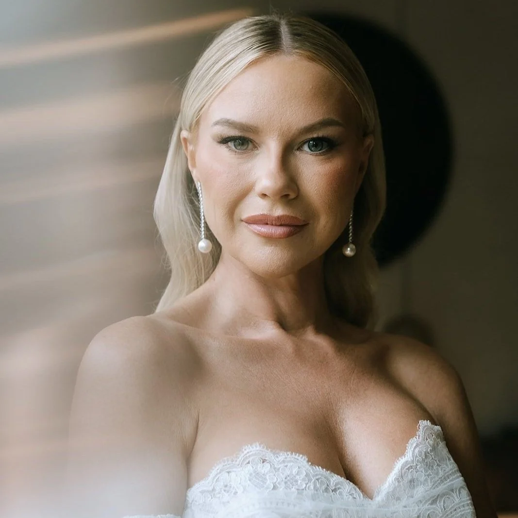 A woman with blonde hair and pearl earrings, wearing a white lace strapless top, gazing into the camera with a neutral expression.