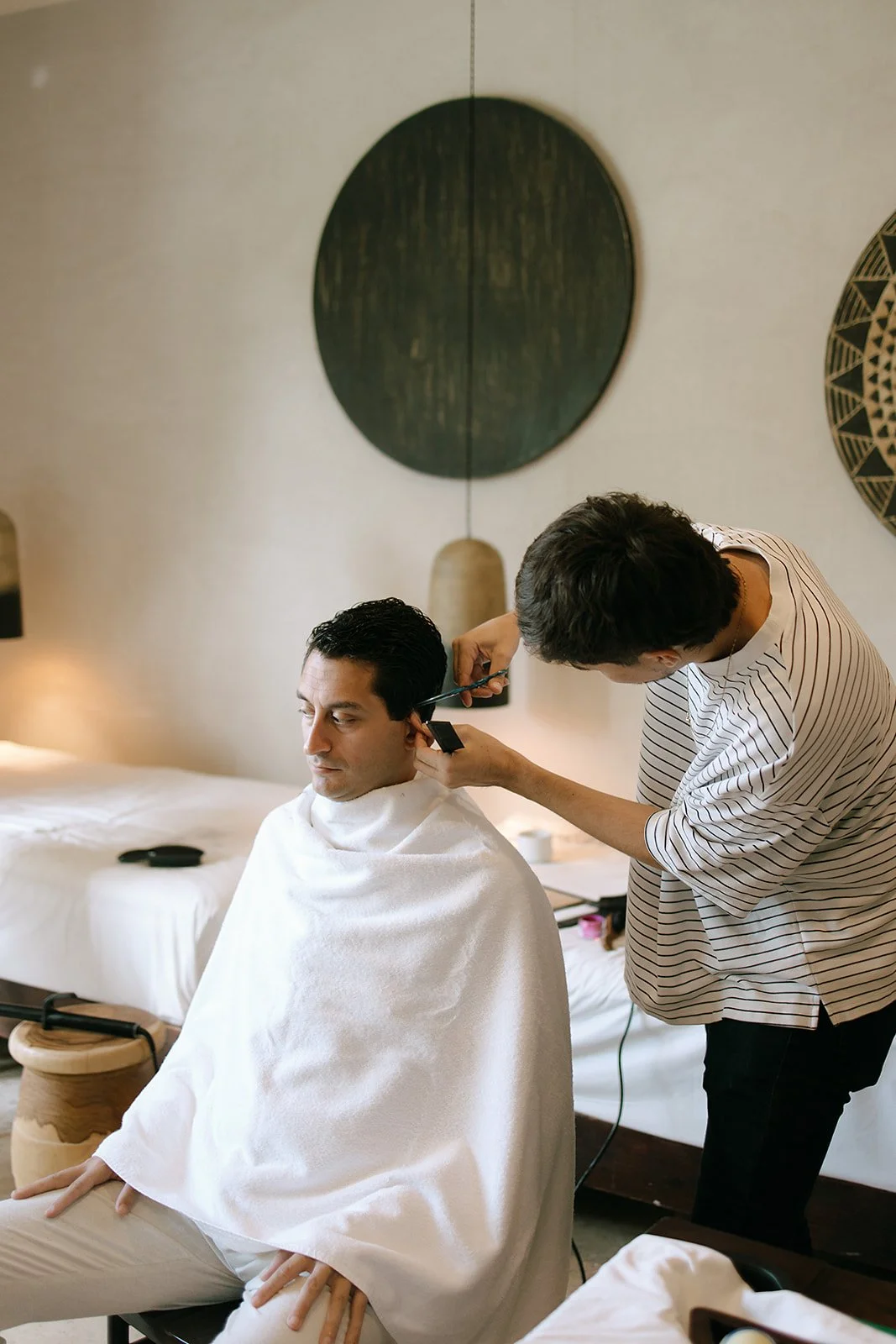 A man getting a haircut in a modern, well-lit room. The barber is focused on cutting the man's hair, who is seated with a white cape. The room decor includes wall art and a hanging pendant light.