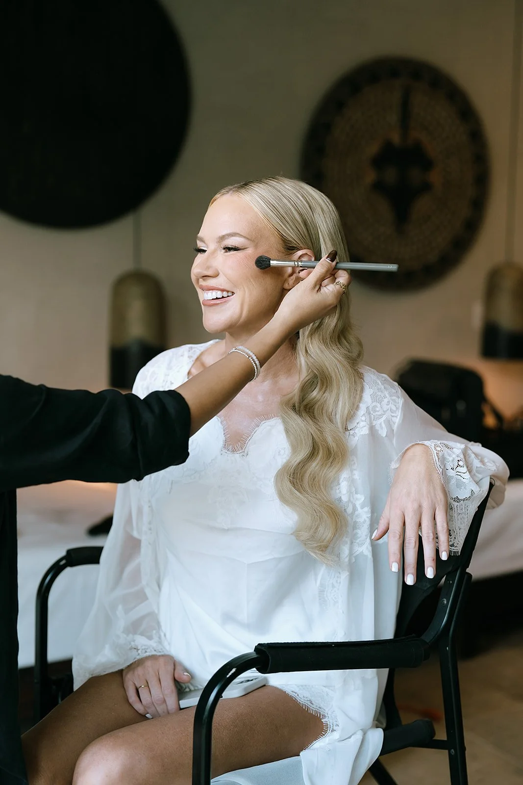 A woman with long wavy blonde hair is sitting in a chair and smiling as a makeup artist applies blush to her cheek using a large makeup brush.