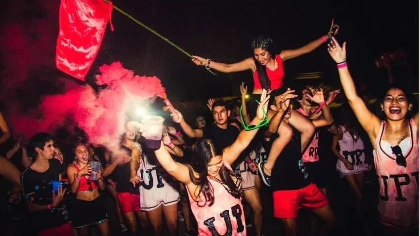 Grupo de jóvenes en una fiesta con luces de bengala y humo rojo, disfrutando y bailando. Una persona sostiene una bandera pequeña mientras otra es llevada en los hombros. Algunos visten camisetas con las letras 'UP'.
