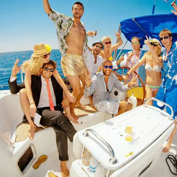Grupo de personas celebrando en un bote, algunas en traje de baño y otras vestidas de manera casual, bajo un cielo despejado y junto al mar.