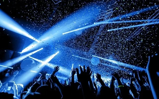 Concierto en vivo con luces azules y confeti, manos levantadas de la multitud disfrutando del evento.