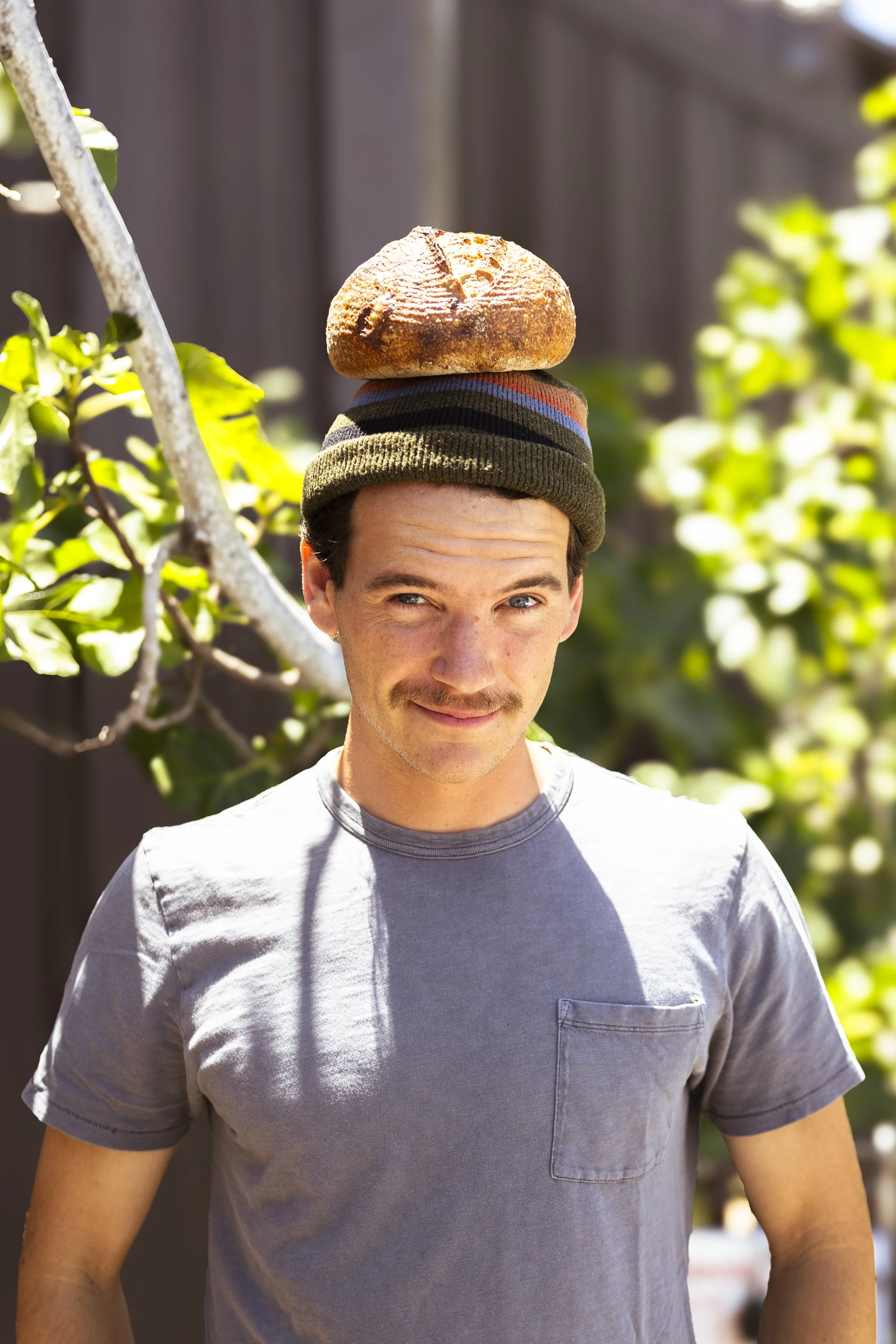 A man wearing a beanie with a loaf of bread on top, standing outdoors with sunlight and greenery in the background.